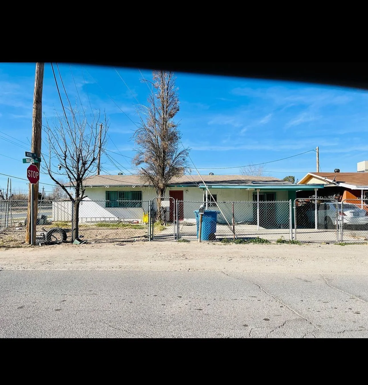 Single-story house with a chain-link fence and a bare tree in front. A stop sign is on the corner, and a blue garbage bin is near the driveway. The sky is clear and blue.