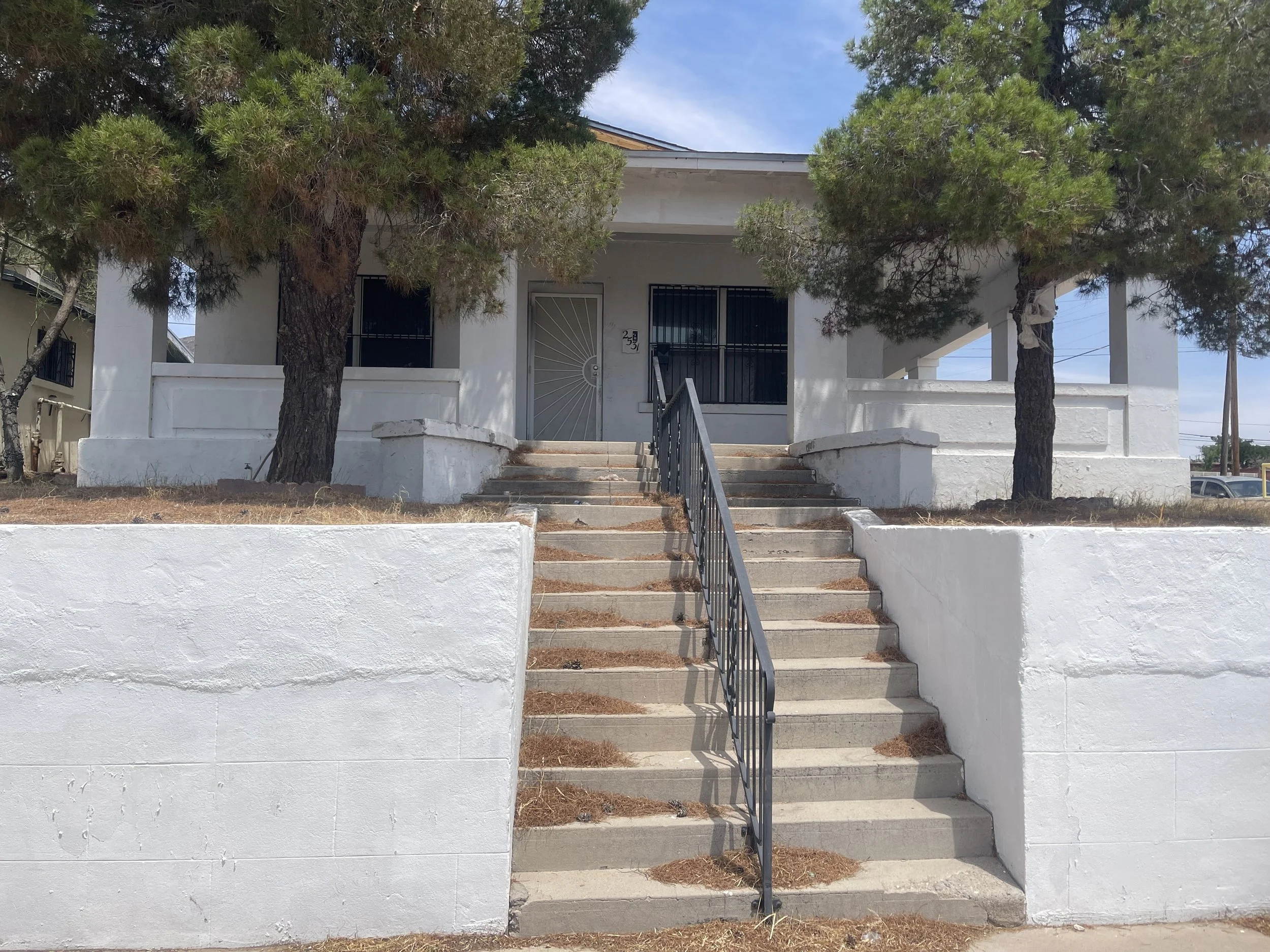 Front view of a white house with a porch, two trees, and a concrete staircase leading up to the entrance.