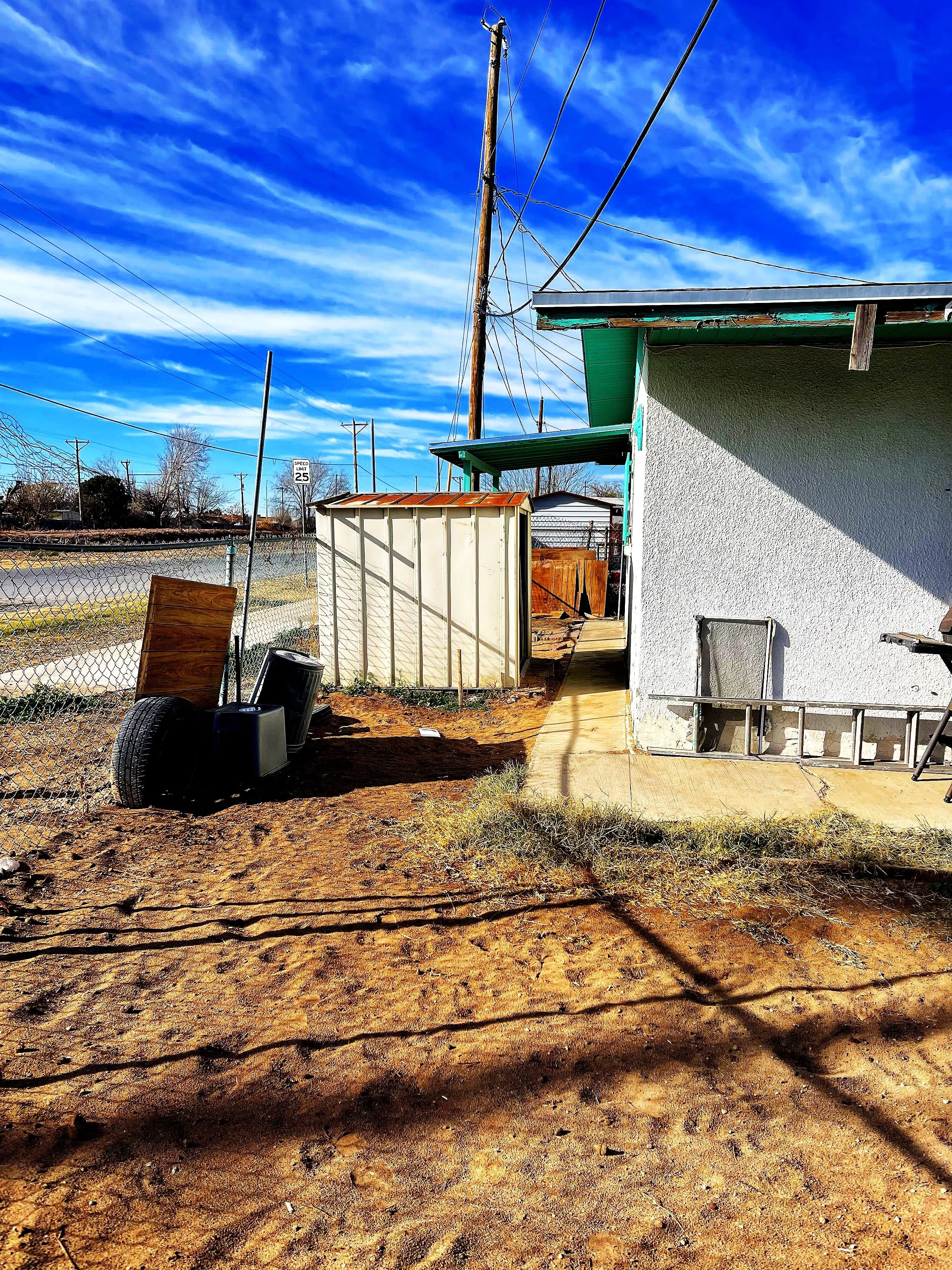 Outdoor urban scene with a rundown building, a chain-link fence, a metal shed, and scattered tires on dirt ground. Utility poles and power lines are visible against a blue sky with wispy clouds.
