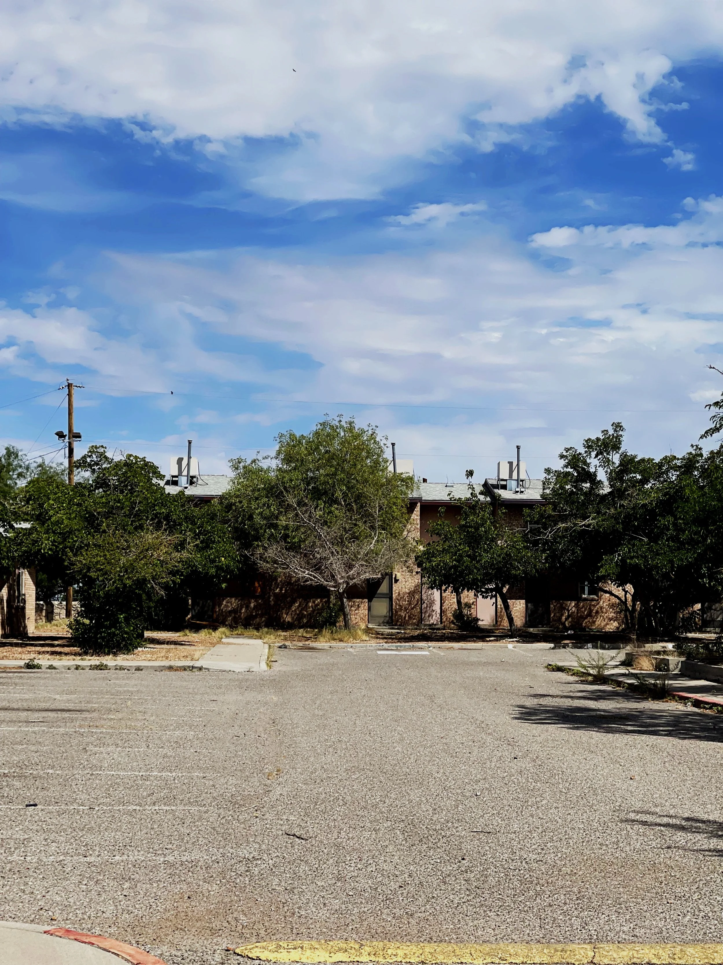 Empty parking lot with faded lines, trees, and a brick building in the background under a cloudy blue sky.