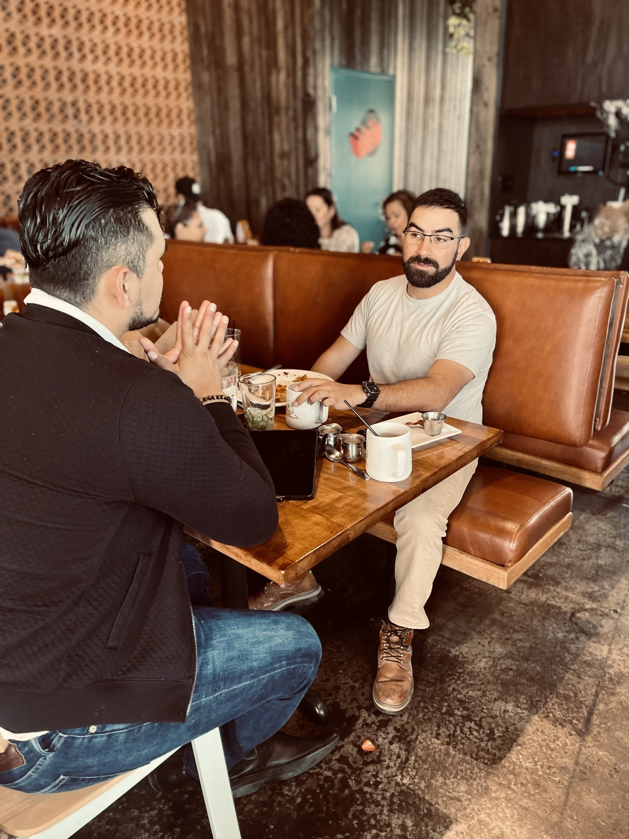 Two men sitting at a wooden table in a cafe, engaging in conversation. One wears a black jacket and jeans, and the other wears a light-colored shirt and pants. Coffee cups and other dining items are on the table.