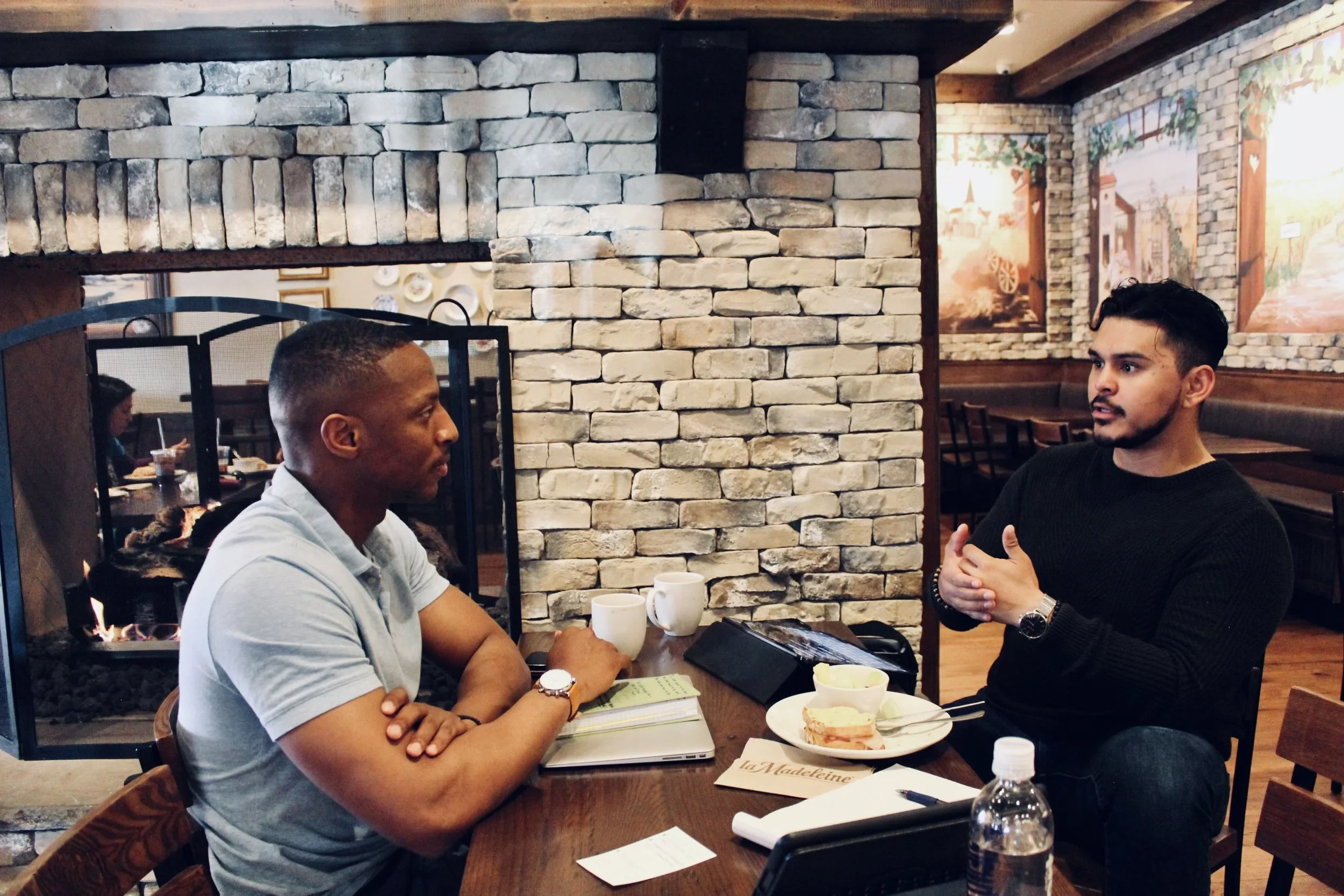 Two men having a discussion at a cafe table, with stone wall and fireplace in the background. They have coffee cups, a sandwich, and a notebook on the table.