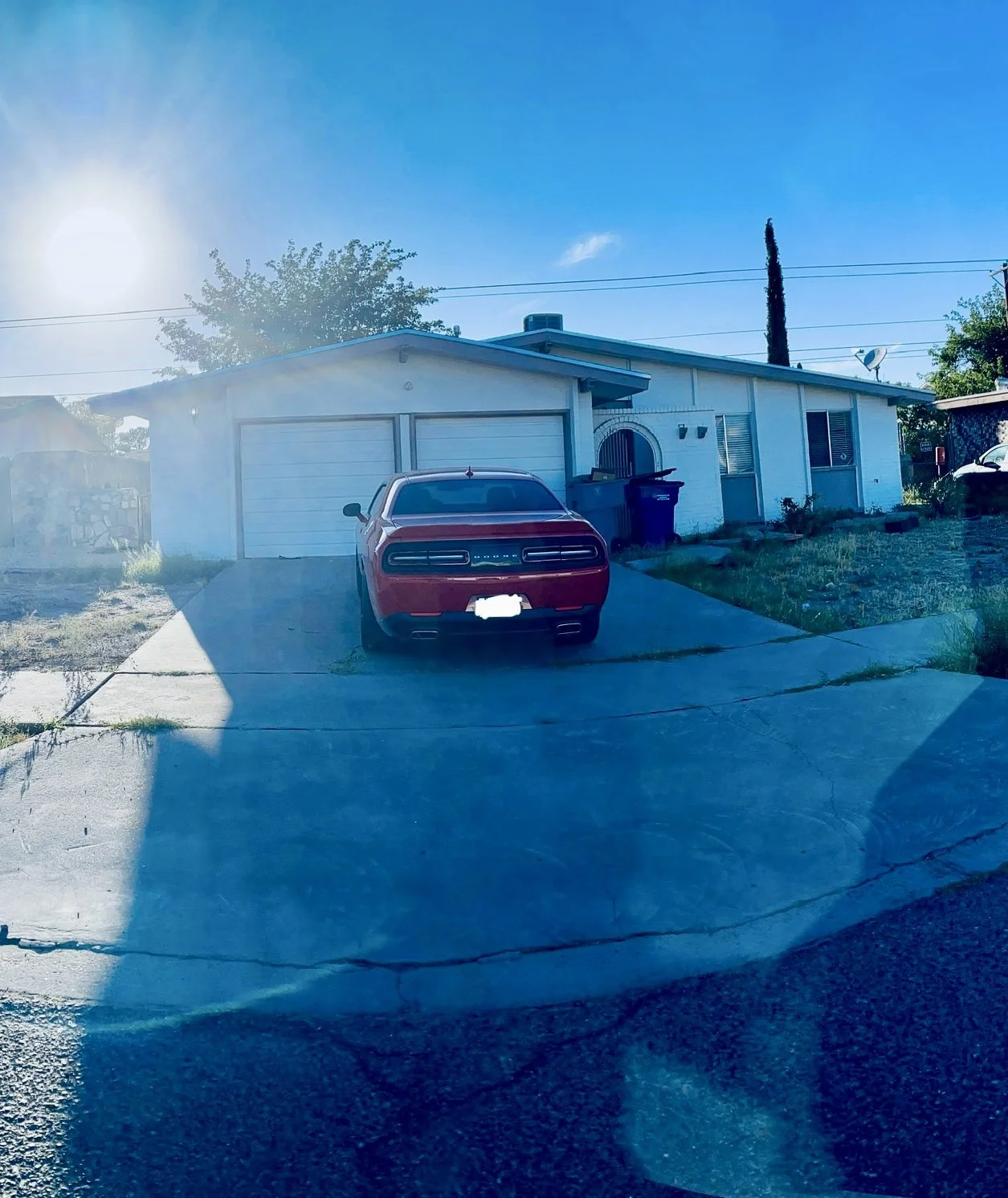 Red car parked in front of a single-story house with a garage and driveway, under a clear blue sky with visible sunlight.