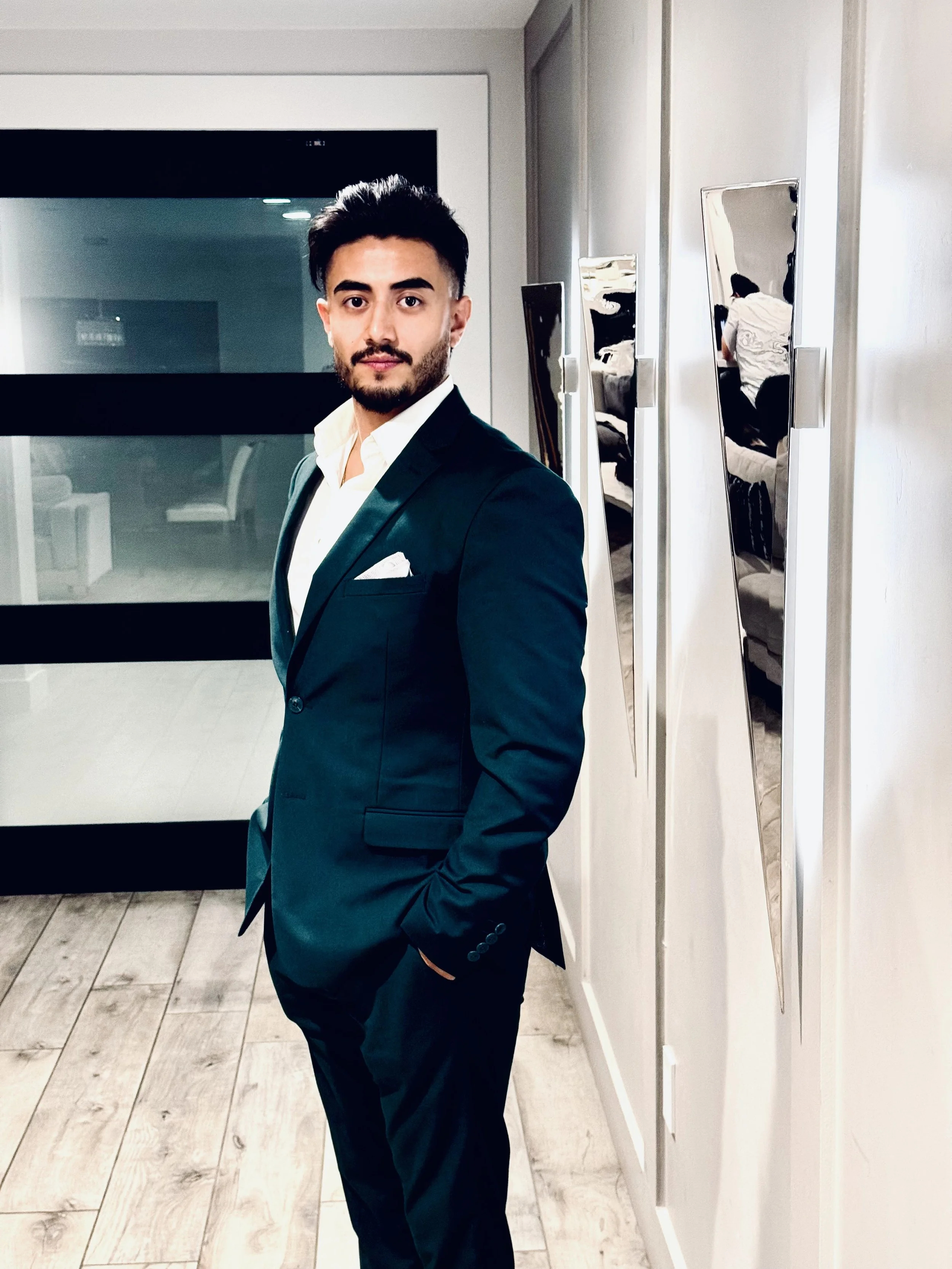 Man in a suit standing in a modern hallway with wood flooring, next to mirrored panels.