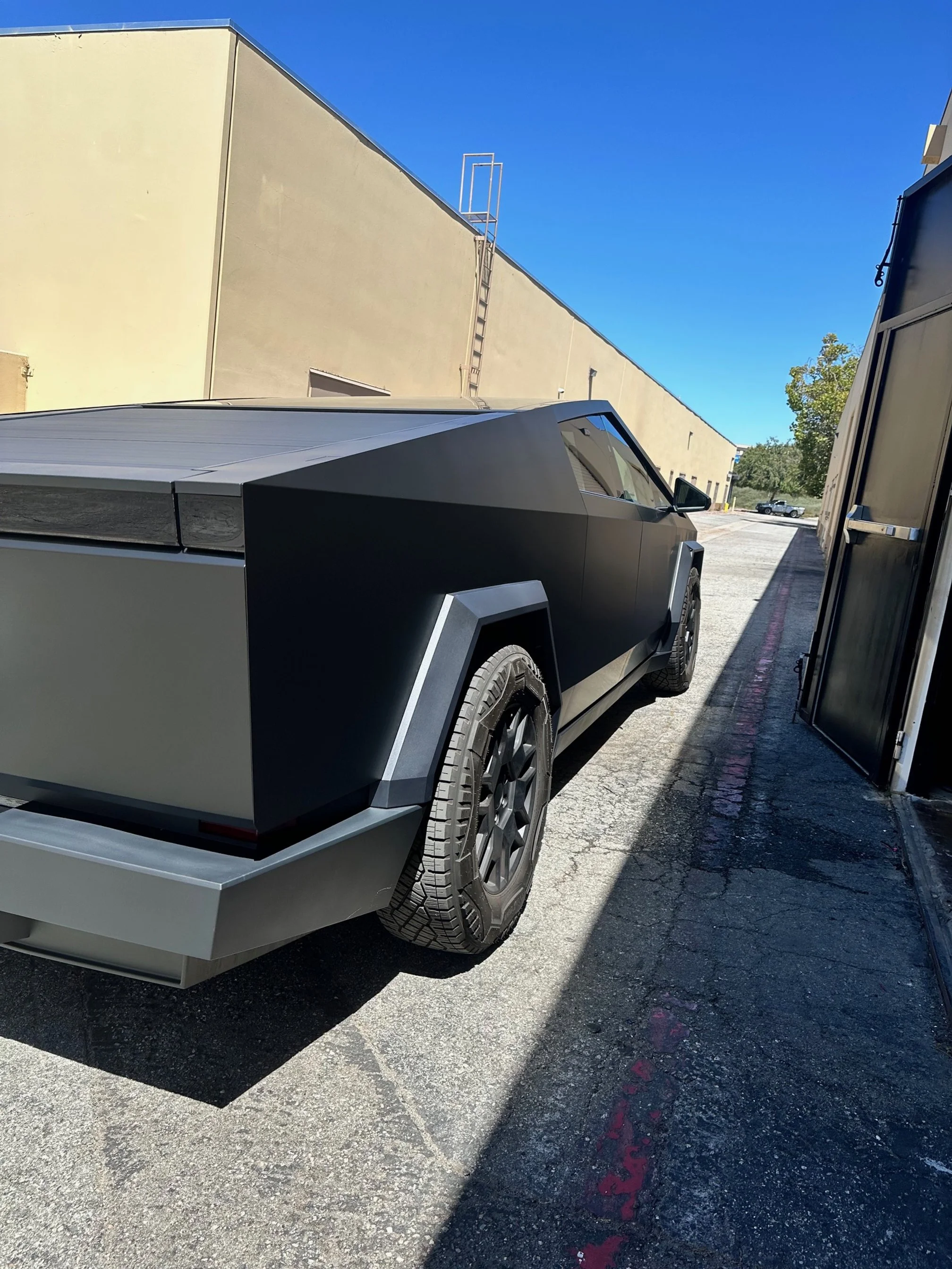 A black, futuristic, angular Tesla Cybertruck parked on a cracked asphalt lot beside a building and storage units with a clear blue sky above.