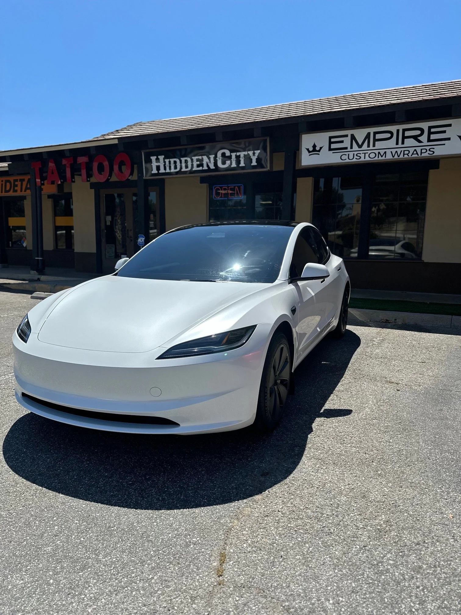 White Tesla parked in front of a storefront with signs for 'Hidden City', 'Empire Custom Wraps', and 'Tattoo' under a clear blue sky.