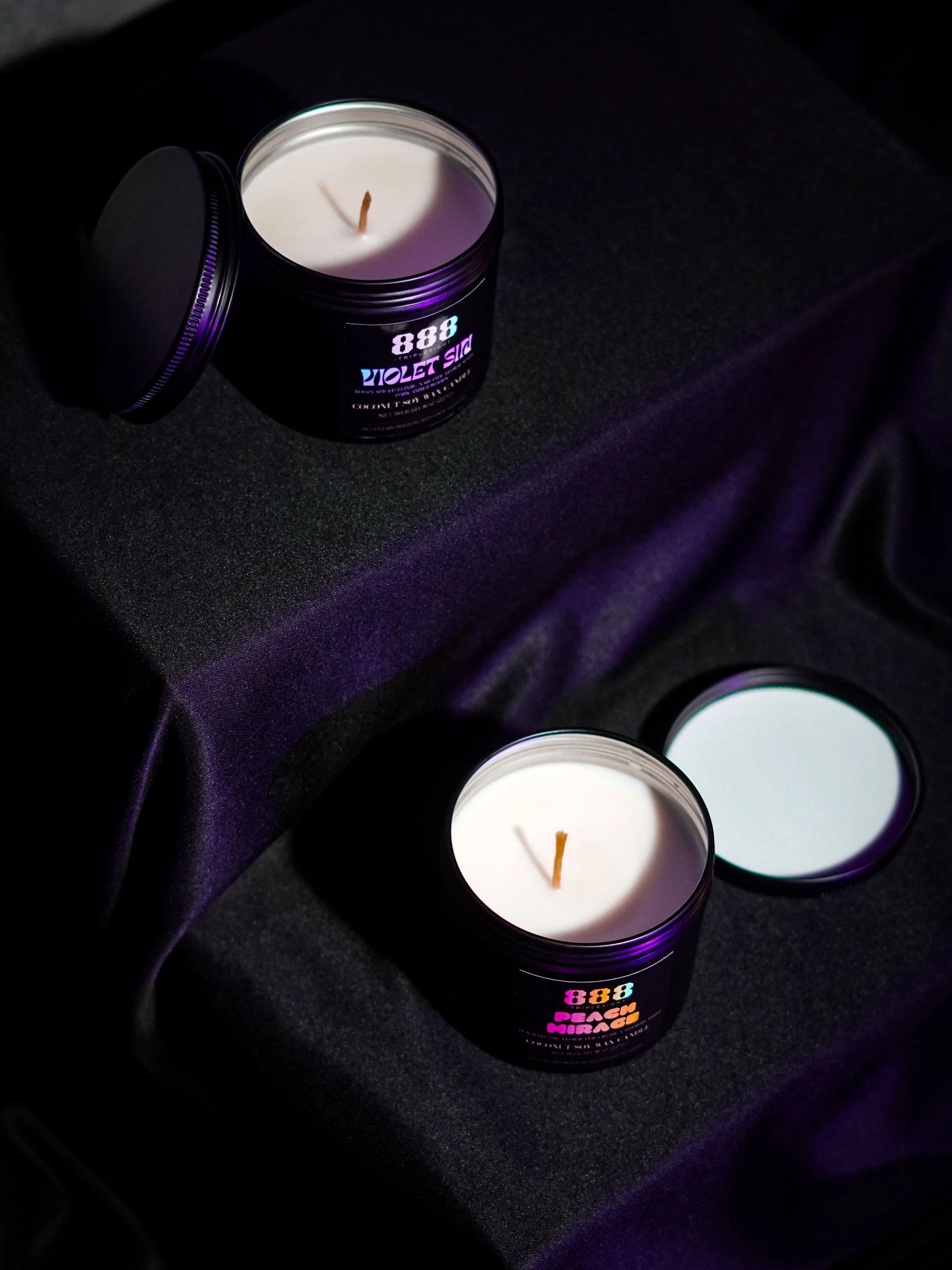 Two white 888 candles in black containers with one lid open, placed on a black surface with purple lighting, and a mirror reflecting the candles. Signature Scent