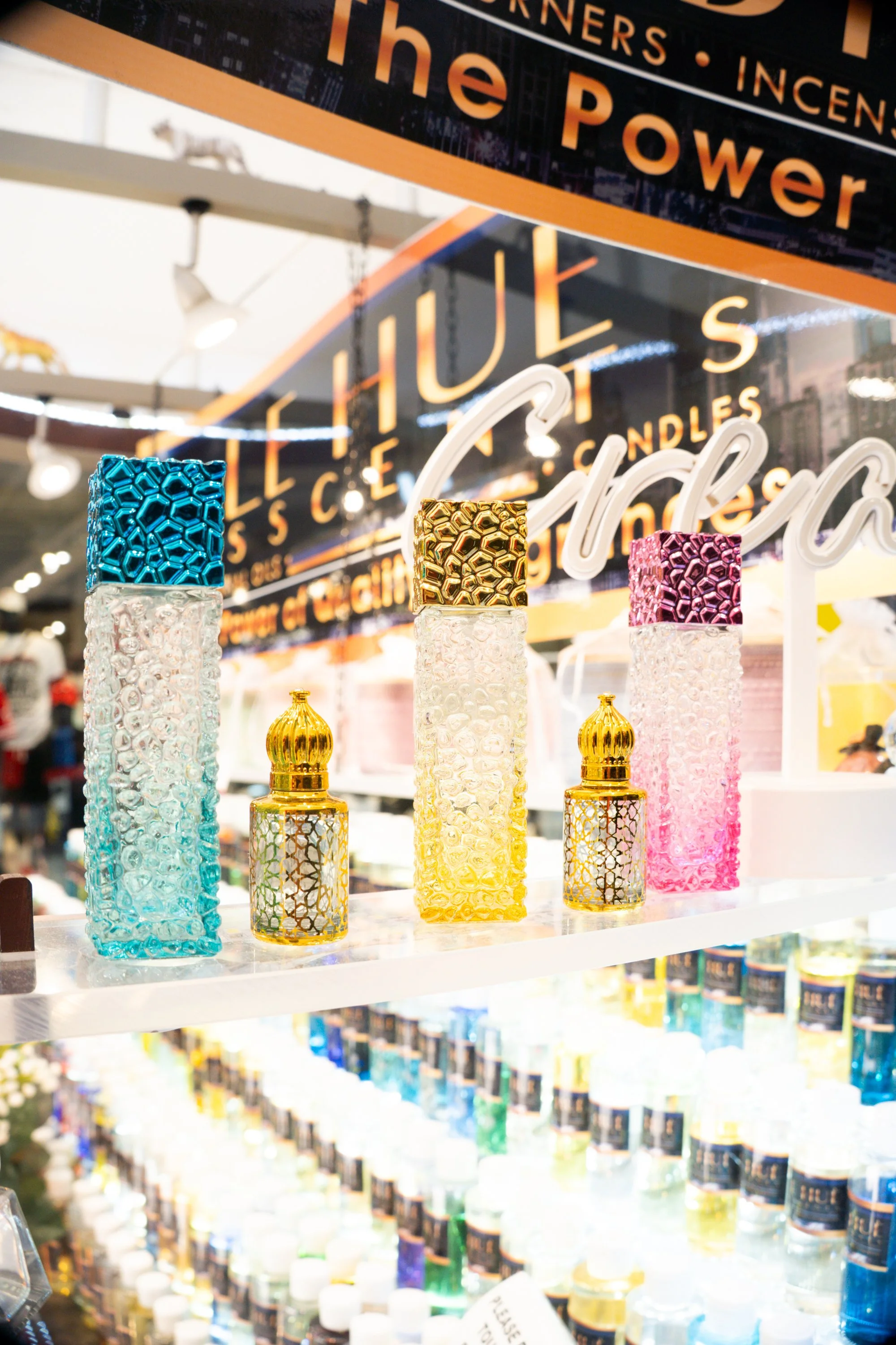 A woman with dark hair, wearing floral accessories, holds a white fan with red and beige floral designs, partially covering her face. She is dressed in colorful clothing and accessories, with a yellow bag and multiple bracelets, sitting outdoors.