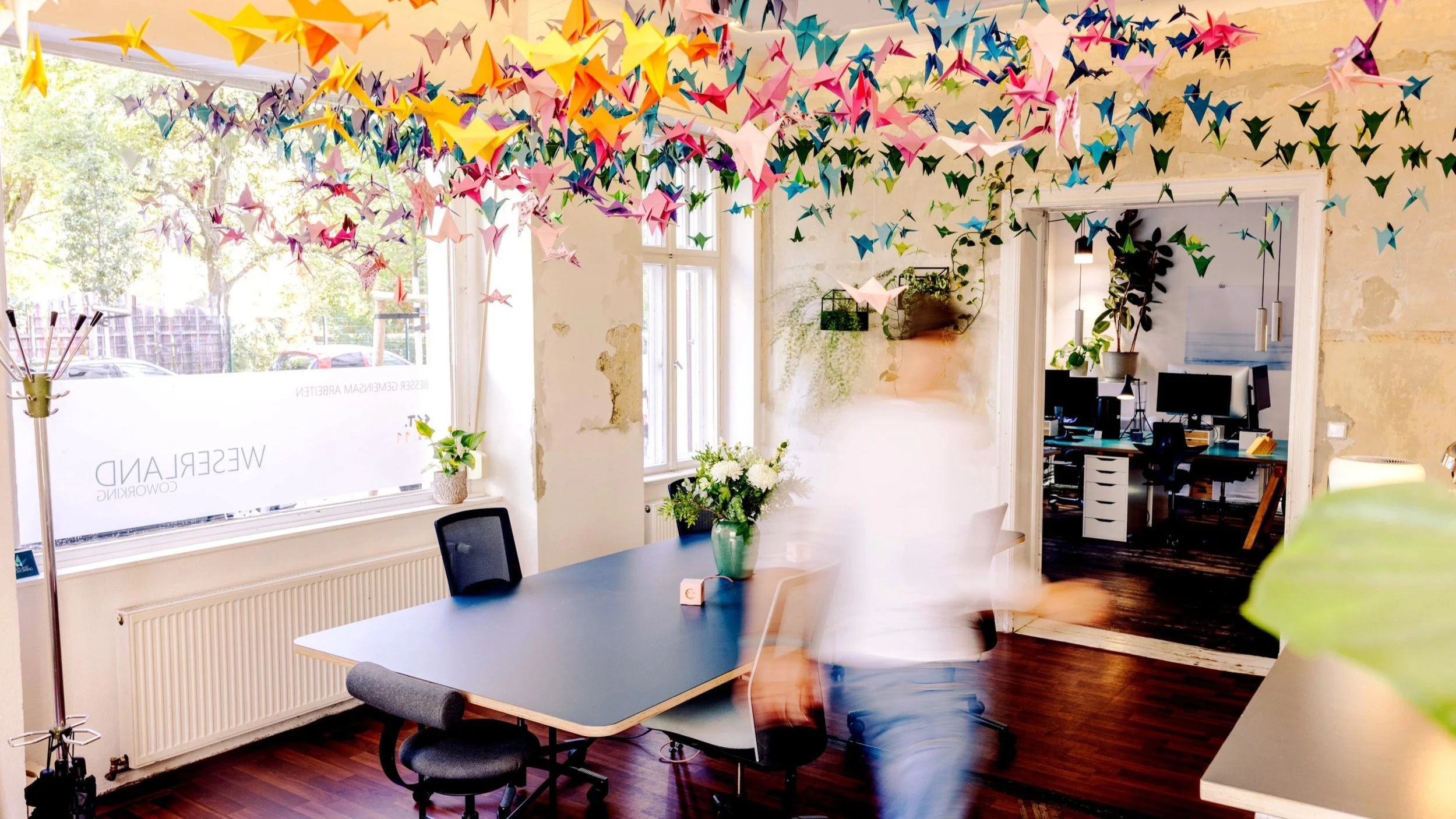 Office with several desks, chairs, plants and a decorative ceiling installation of colorful paper cranes.