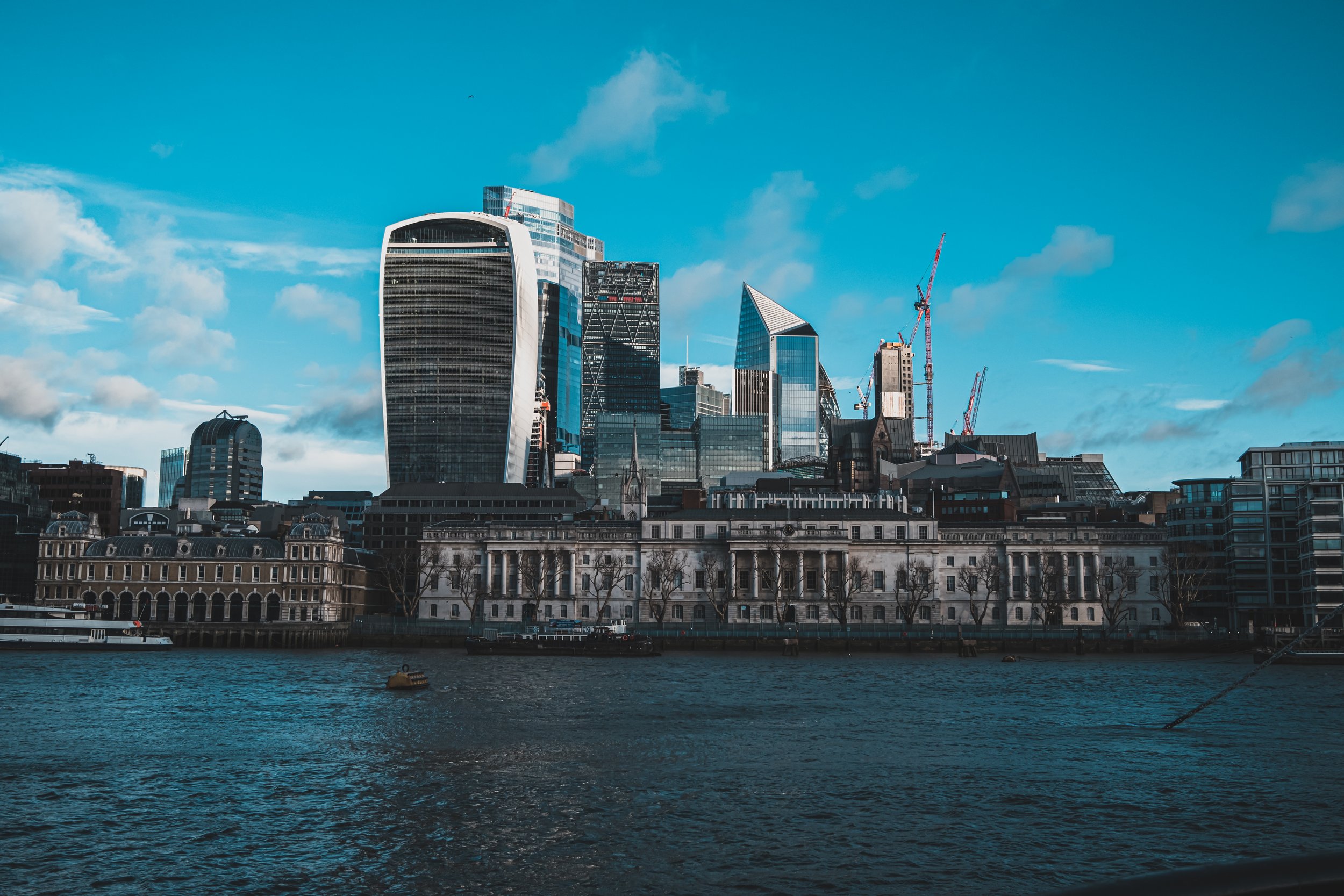 London city skyline with modern skyscrapers and historic buildings along the river Thames under a blue sky with scattered clouds.