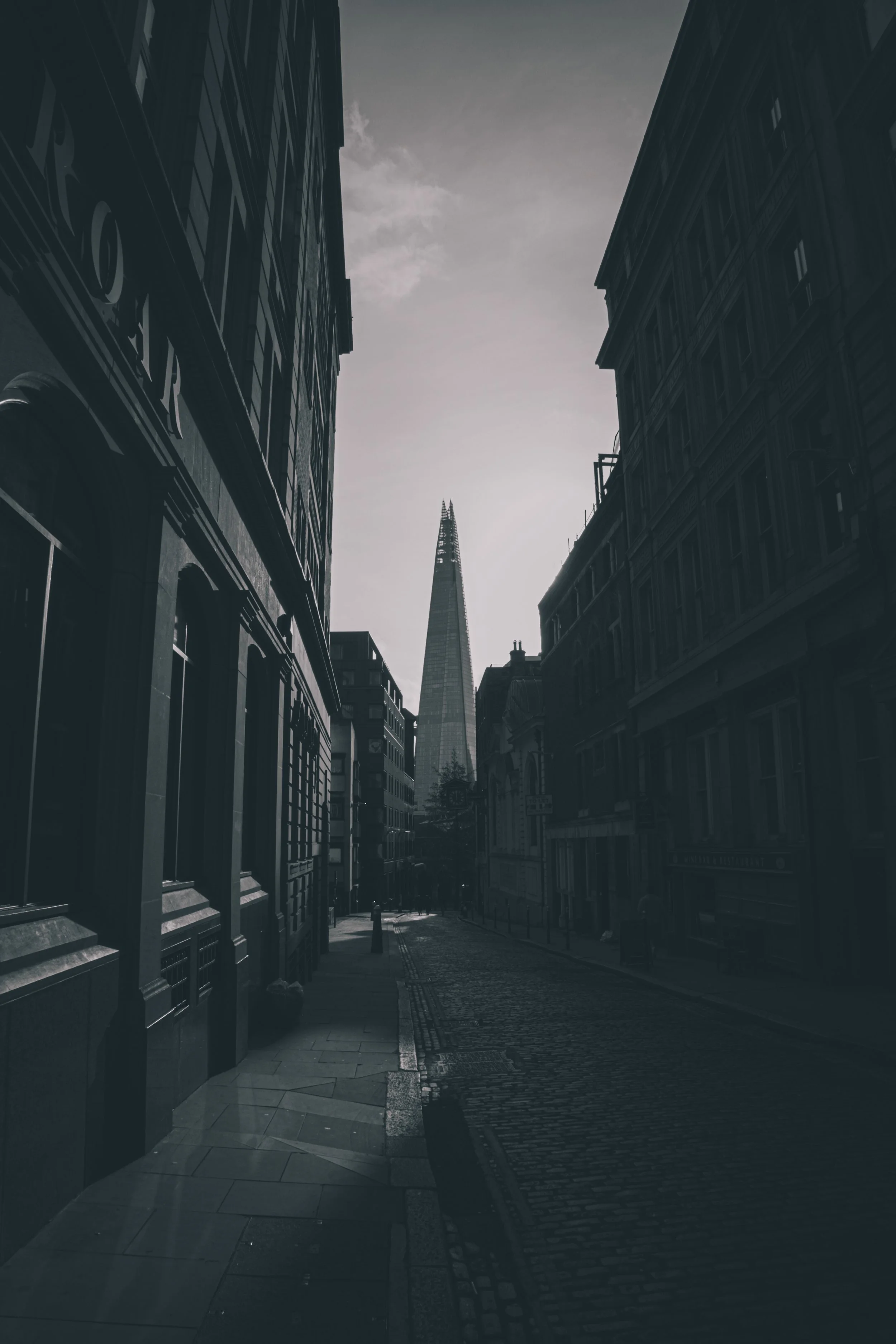 A narrow city street with tall buildings on either side, leading toward the iconic Shard skyscraper in London, with cloudy sky overhead.