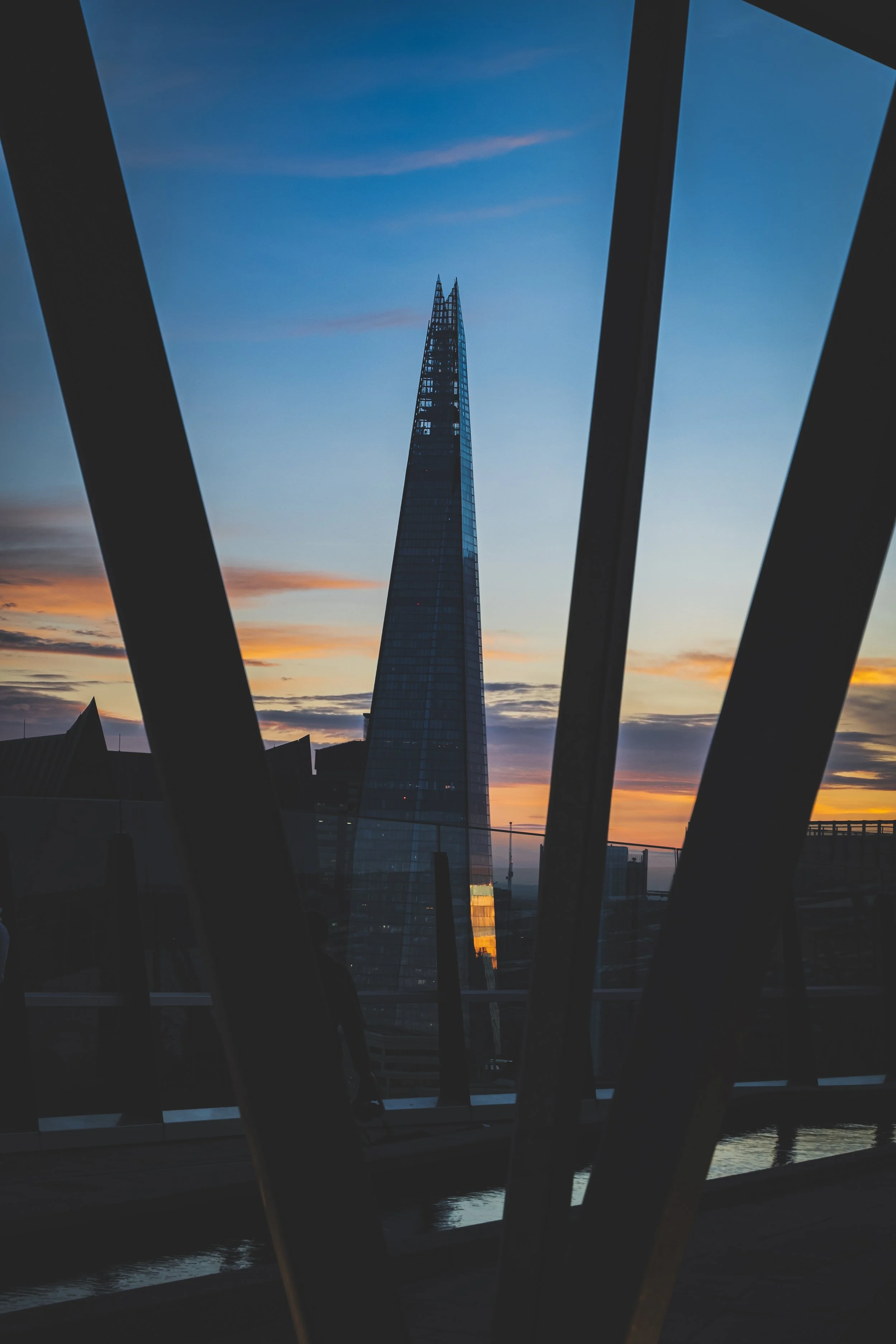 A photograph of The Shard skyscraper taken from a nearby viewing area during sunset, with metal framing in the foreground.