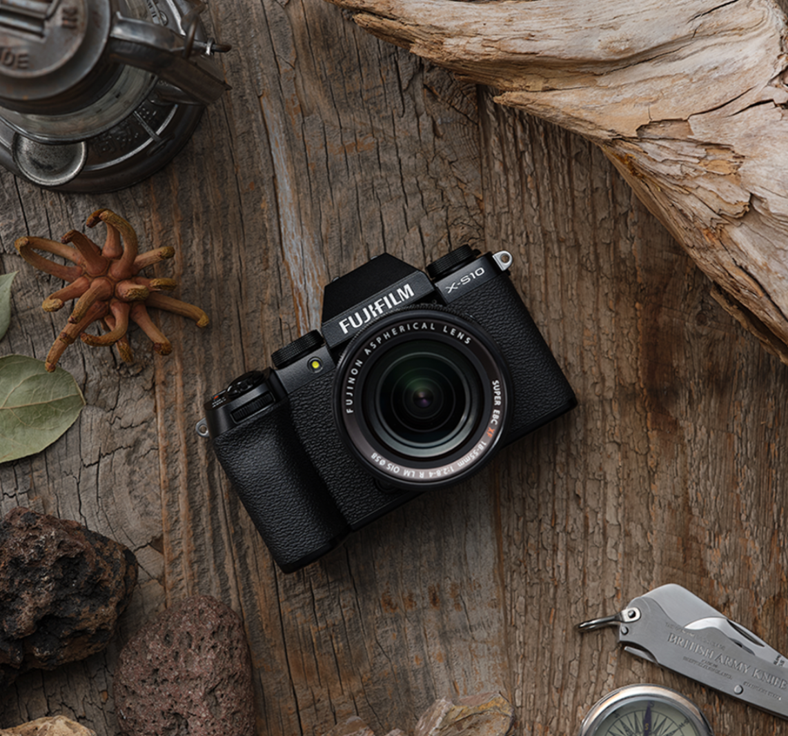 A Fujifilm X-S10 camera with a Fujinon lens on a rustic wooden surface surrounded by rocks, a dried plant, a leaf, a pocket knife, a compass, and an old oil lantern.