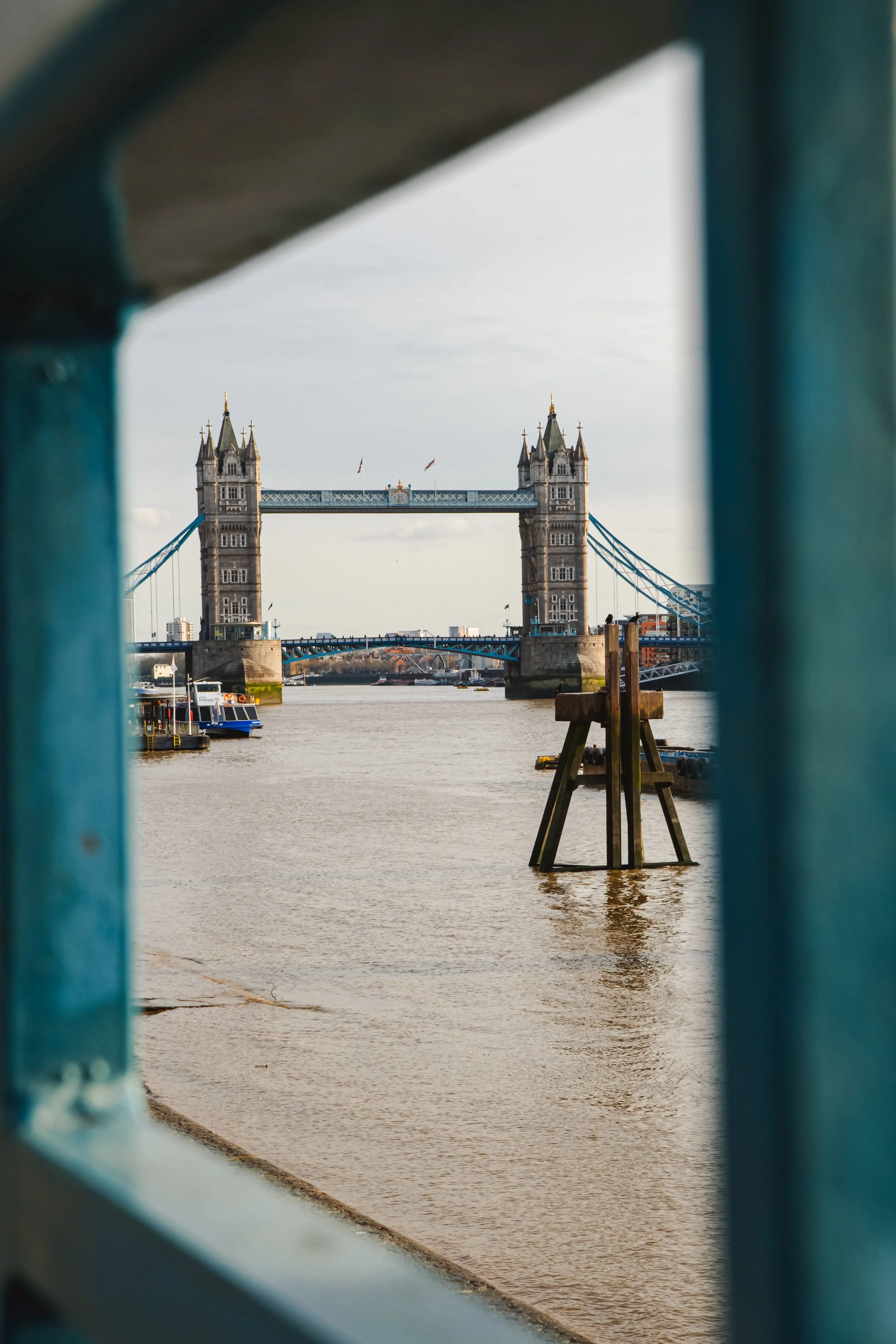 View of Tower Bridge in London through an opening in a blue railing, showing the bridge's towers and suspension cables over the River Thames.