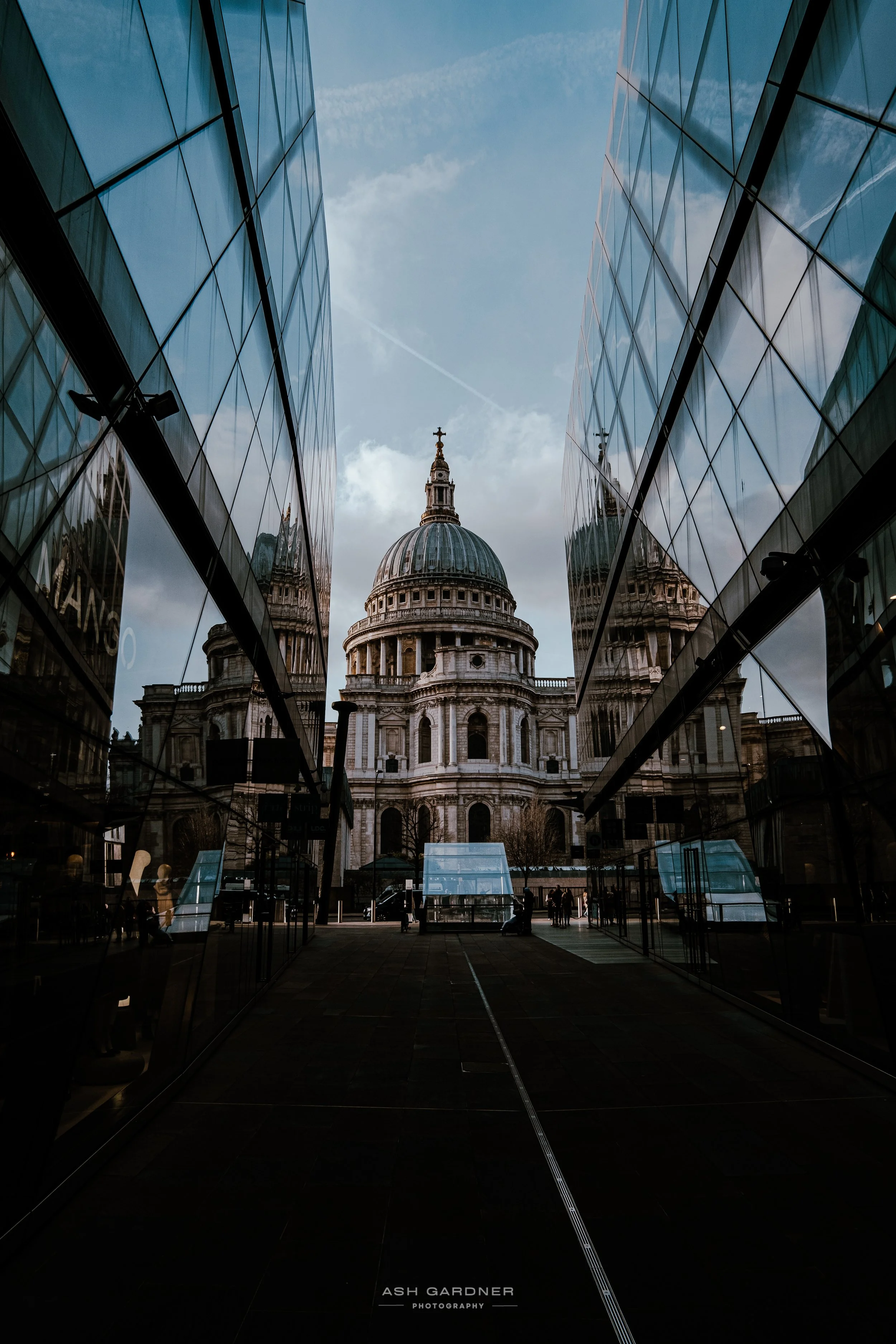 The image shows St. Paul's Cathedral in London, viewed through the reflective glass walls of a modern building. The sky is partly cloudy with a contrail visible, and people are walking around the area.