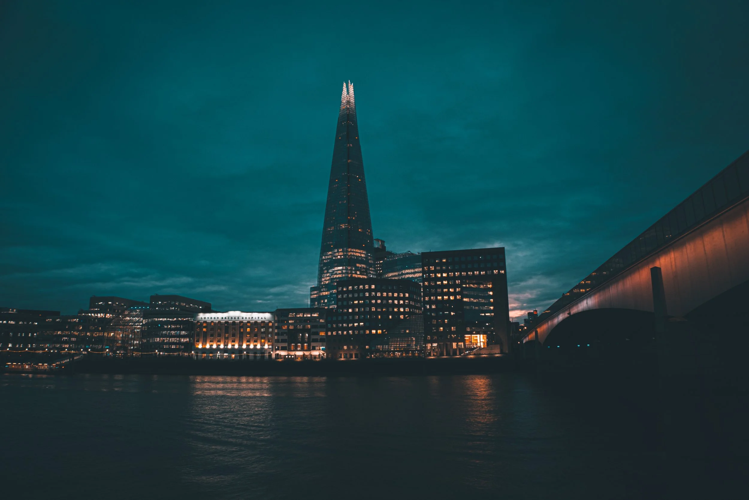 Nighttime cityscape of London featuring The Shard skyscraper illuminated against a dark cloudy sky, with reflections on the river below and a bridge on the right.