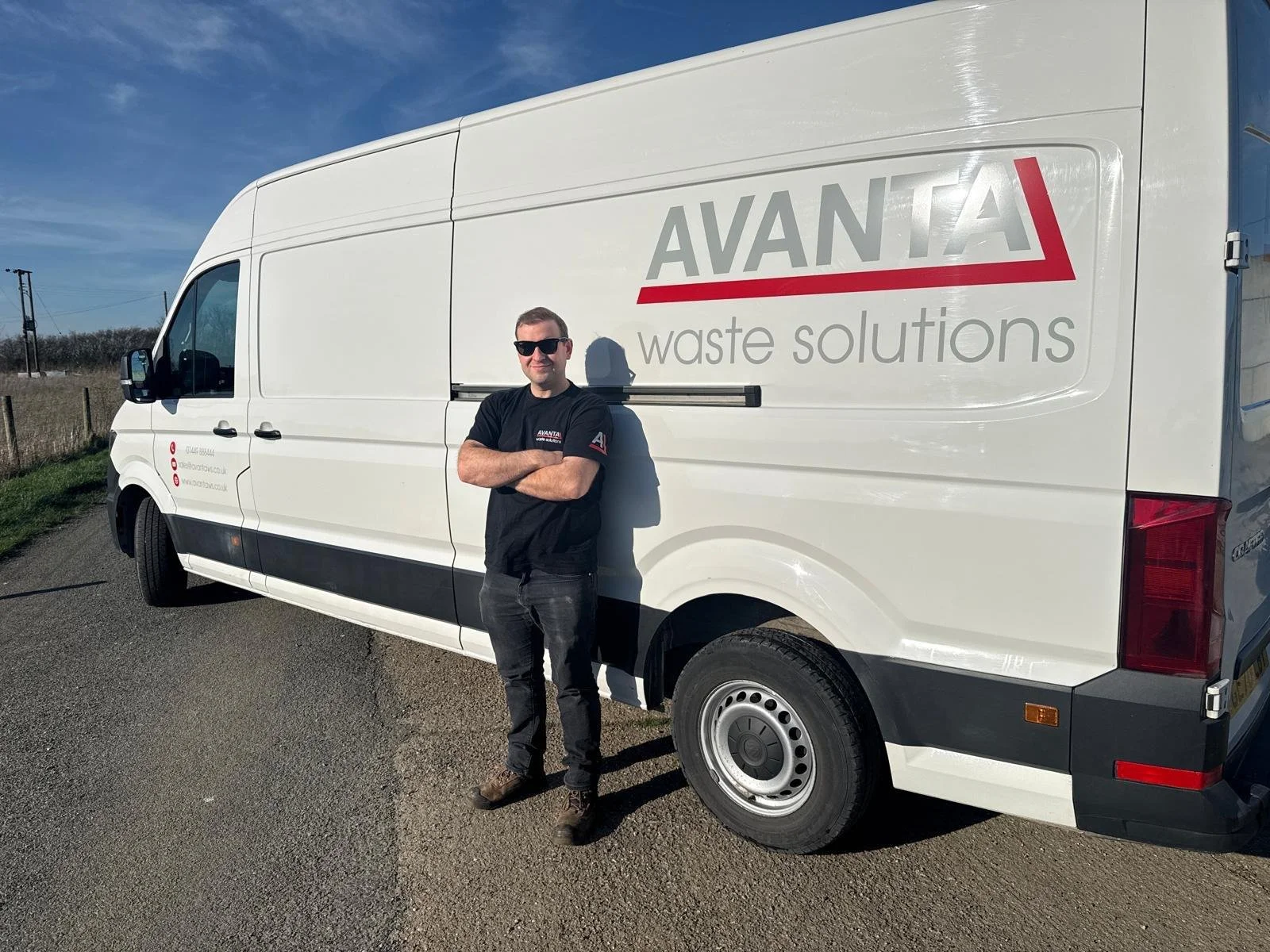 A man in black sunglasses and a black T-shirt with 'Avanta Waste Solutions' logo stands with arms crossed next to a white waste services van with similar branding, parked on a rural road under a blue sky.