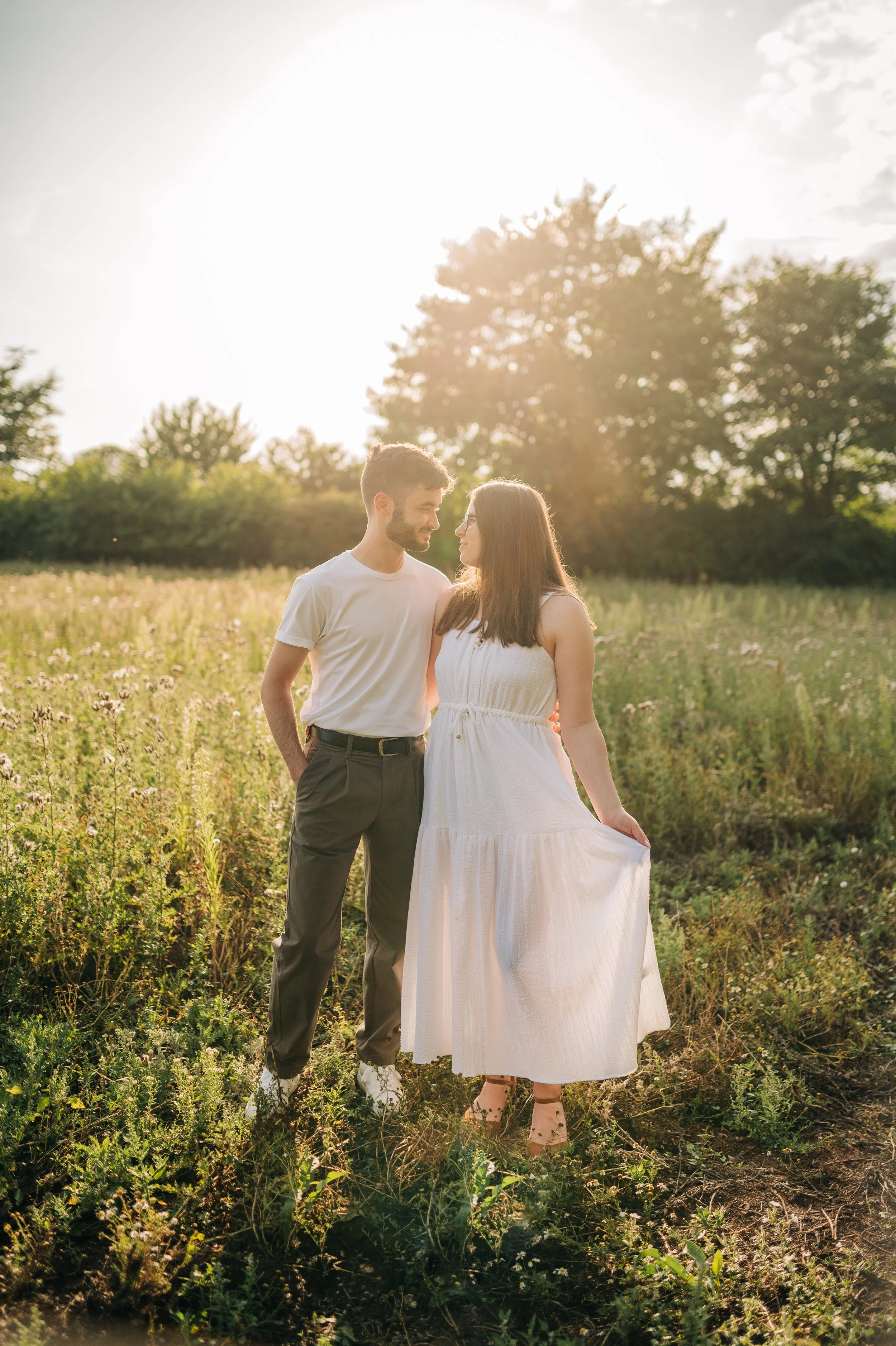Norfolk Wedding Photographer.  A young couple standing in a grassy field with trees in the background, holding hands and gazing at each other during sunset.