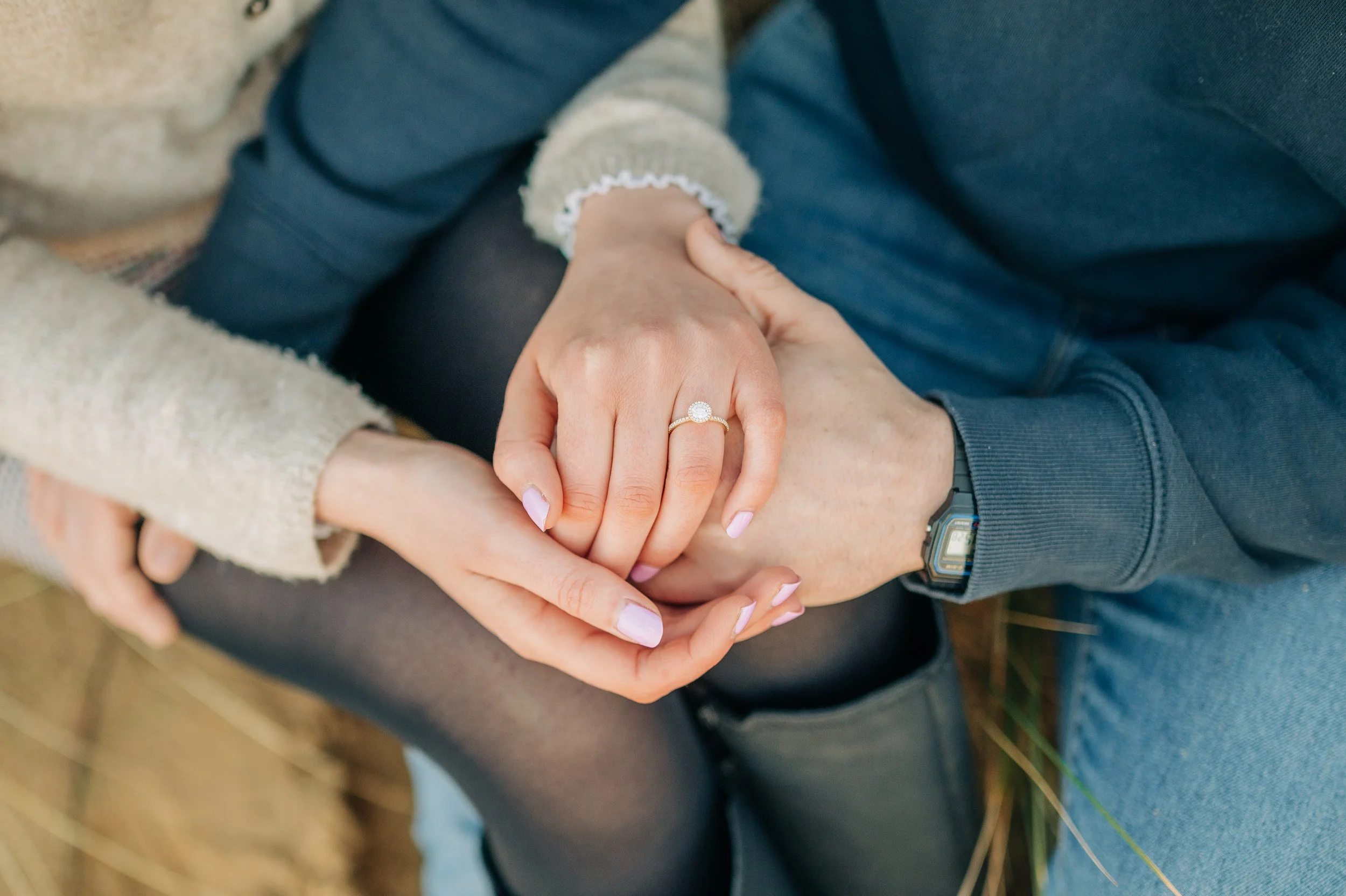 Norfolk Wedding Photographer.  Close-up of a man and woman holding hands, with the woman's hand displaying an engagement ring. The woman is wearing a cozy sweater, and the man is in a dark jacket and a watch.