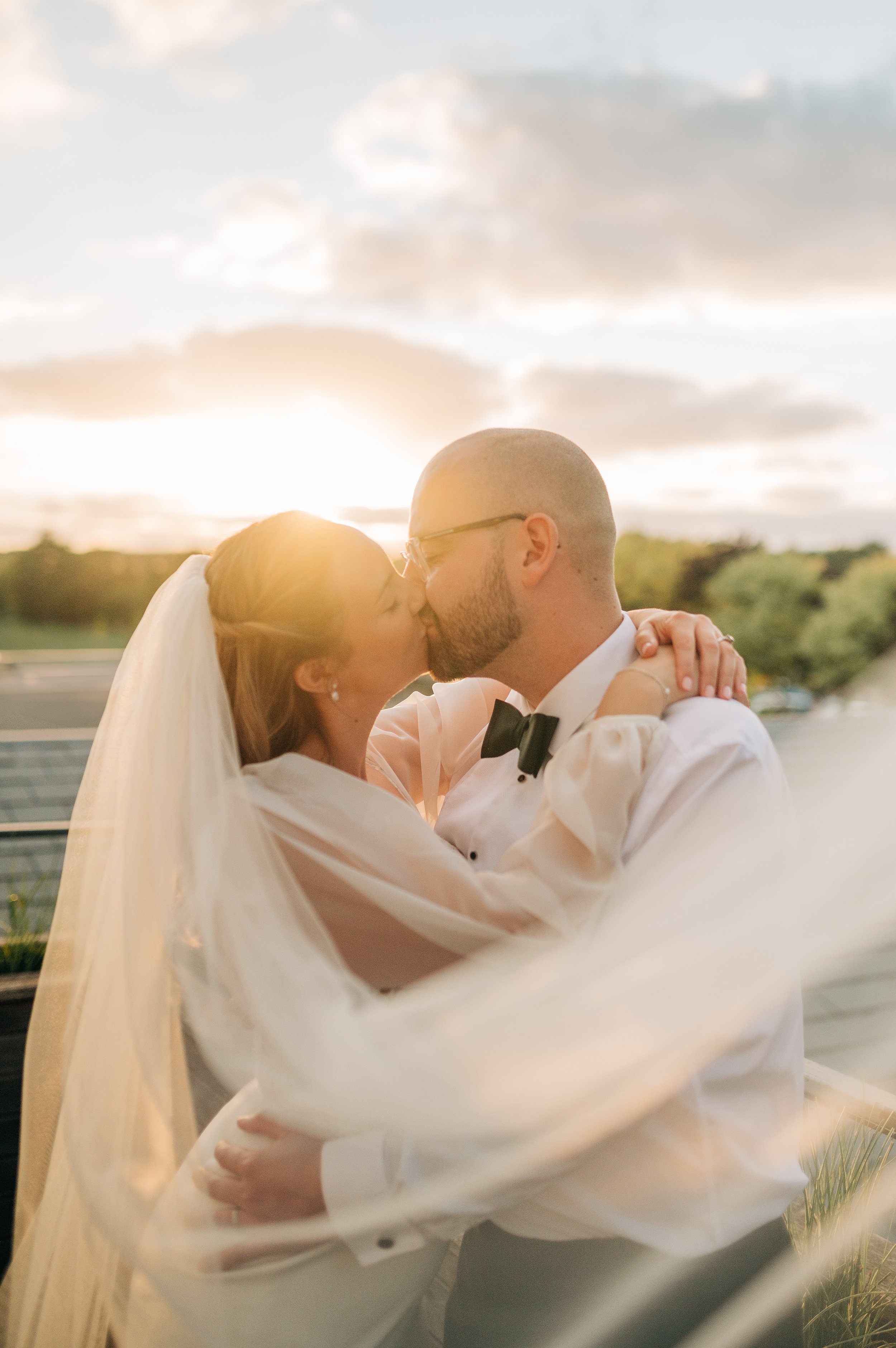 Norfolk Wedding Photographer. A bride and groom kissing outdoors at sunset, dressed in wedding attire. The bride is wearing a veil and earrings, and the groom is wearing glasses and a tuxedo with a bow tie.