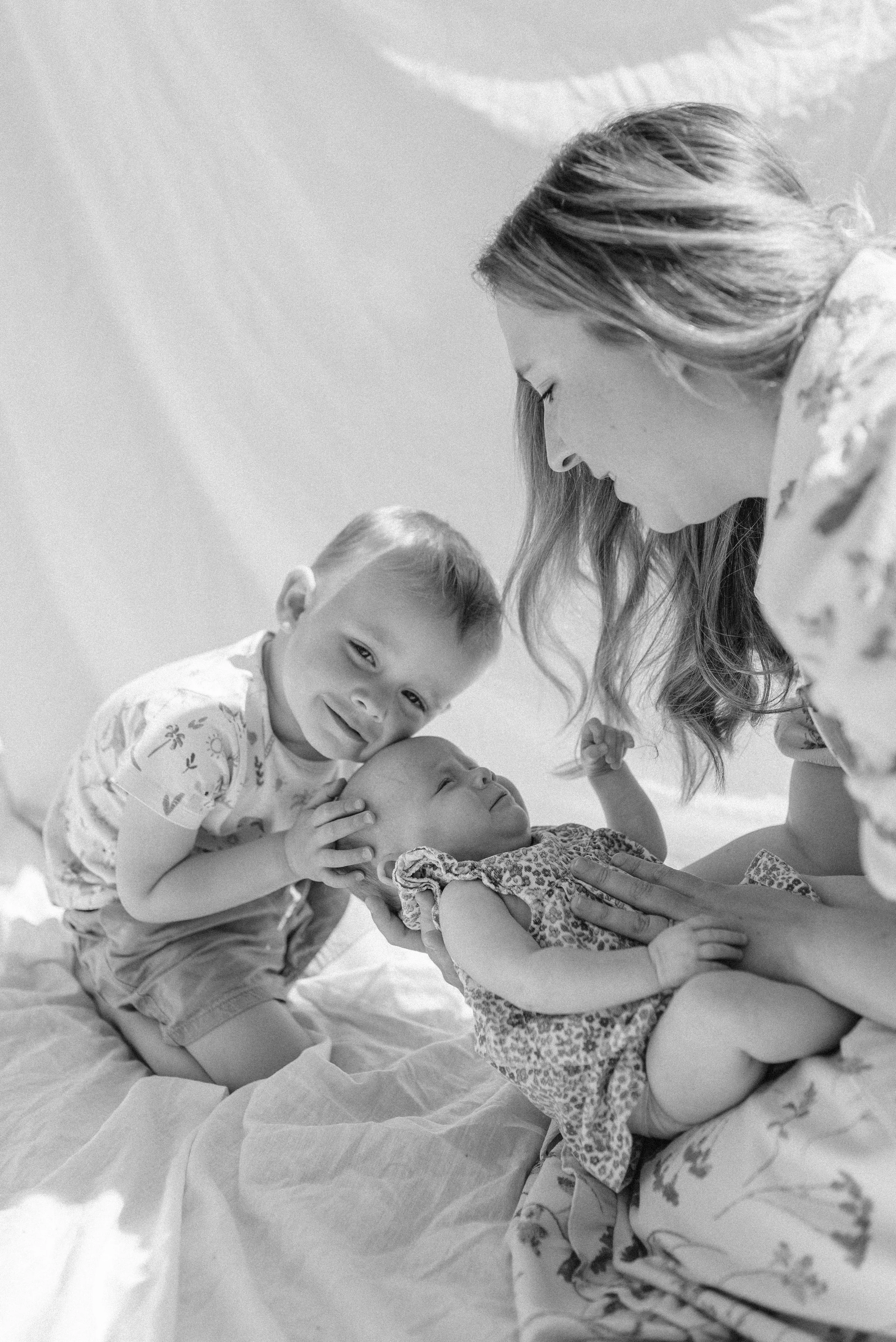 Norfolk Family Photographer.  A woman with long hair leaning over a baby lying on a blanket, with a young boy beside them gently holding the baby's head. The boy is smiling at the camera while the woman looks at the baby.