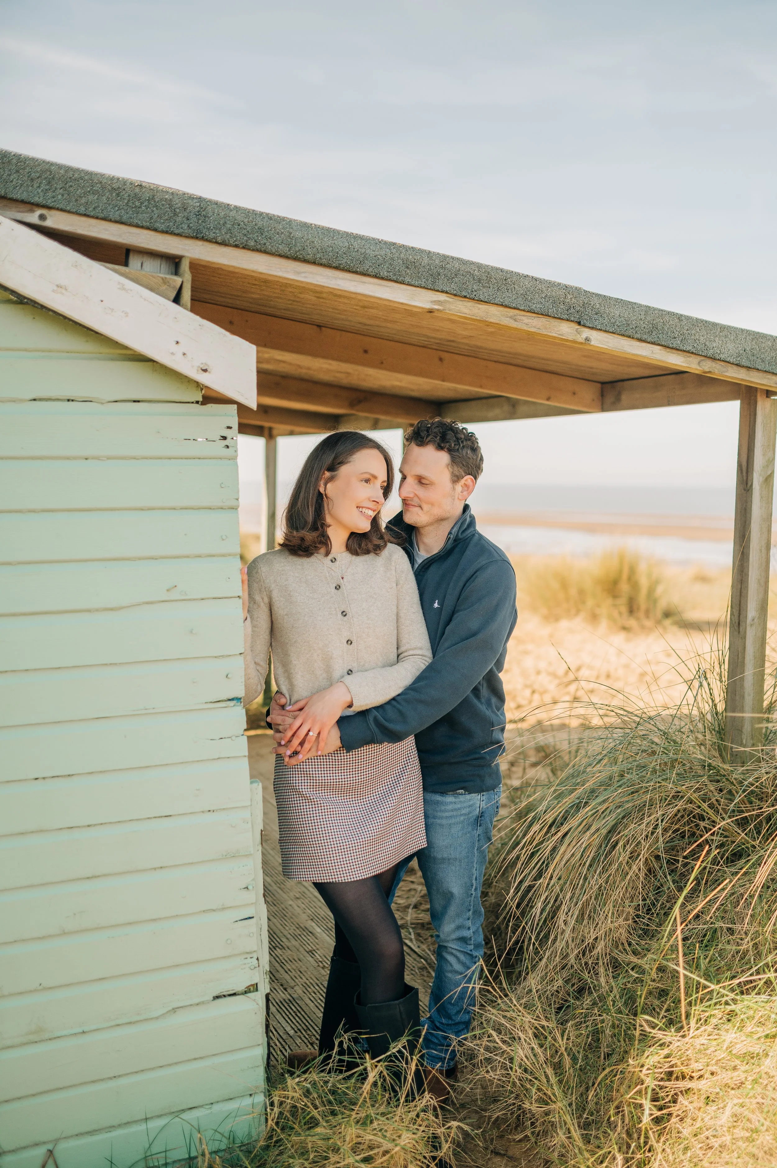 Norfolk Wedding Photographer.  A couple stands close together at a beach hut, smiling and looking at each other, with the ocean and sand dunes in the background.
