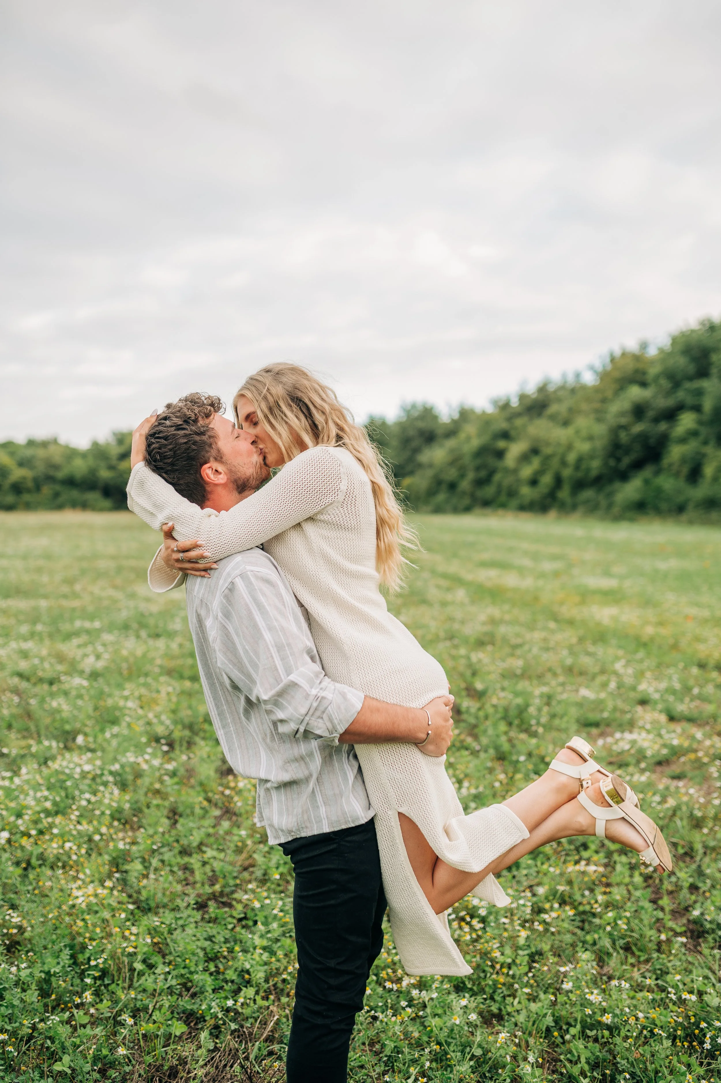 Norfolk Wedding Photographer.  A couple stands in a grassy field, with the man lifting the woman in his arms as they share a kiss. The woman is wearing a white dress and sandals, and the man is dressed in a striped shirt and dark pants.