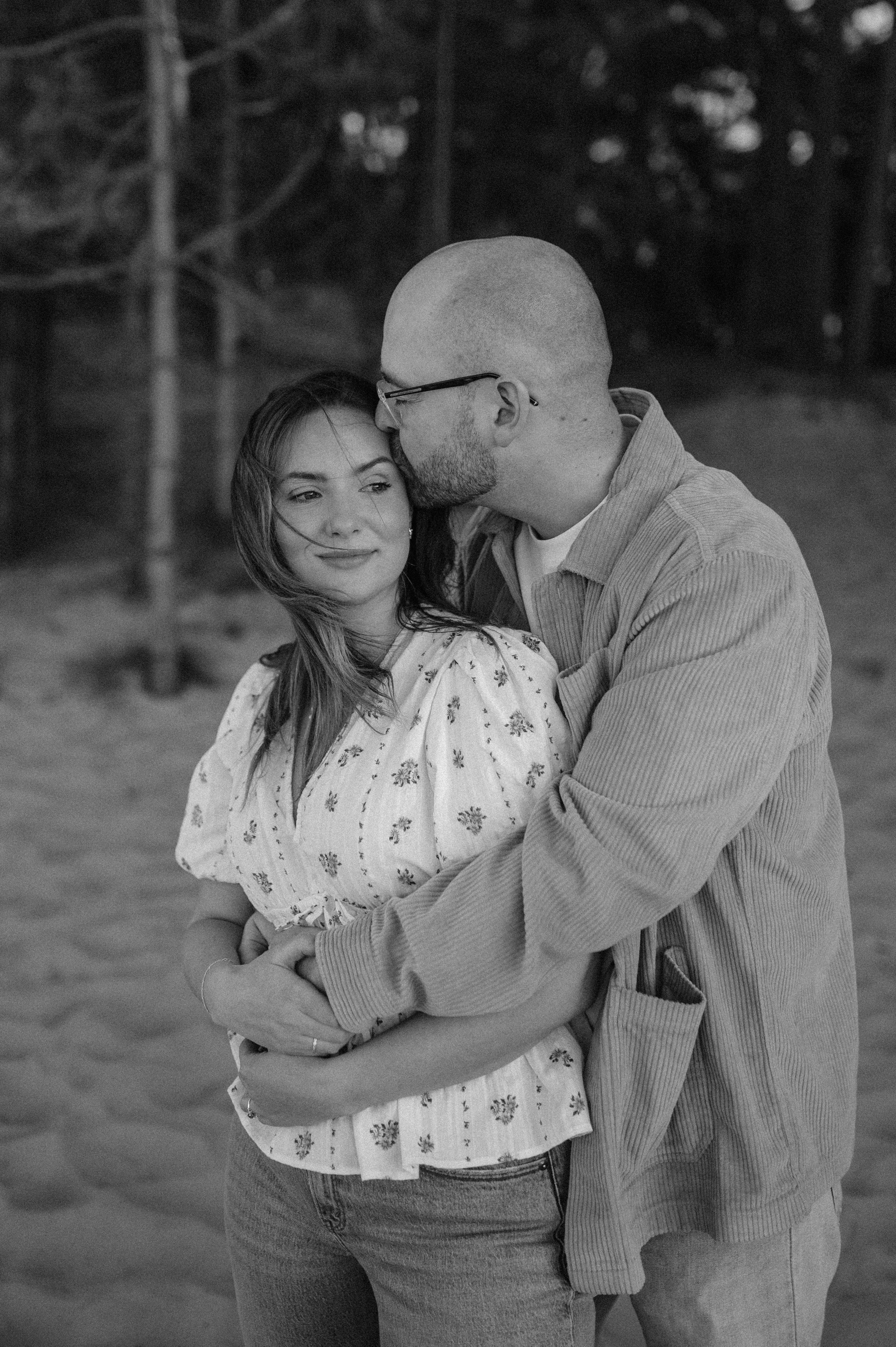 Norfolk Wedding Photographer.  A couple embracing outdoors, with the man kissing the woman's forehead. She is smiling softly, and they are surrounded by trees in the background.