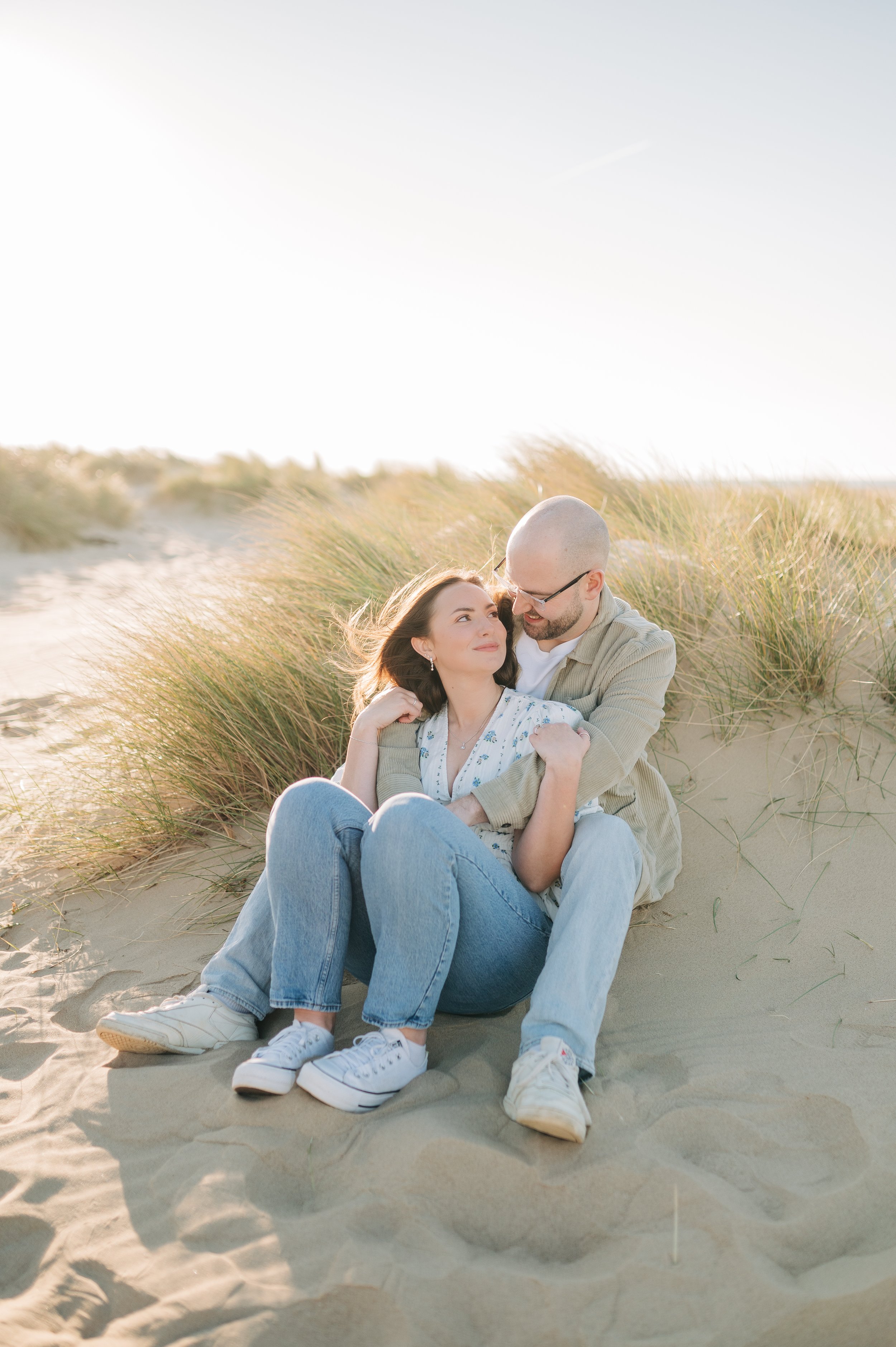 Norfolk Wedding Photographer.  A couple sitting on the sand at a beach, surrounded by tall grass, enjoying a moment together during sunset.