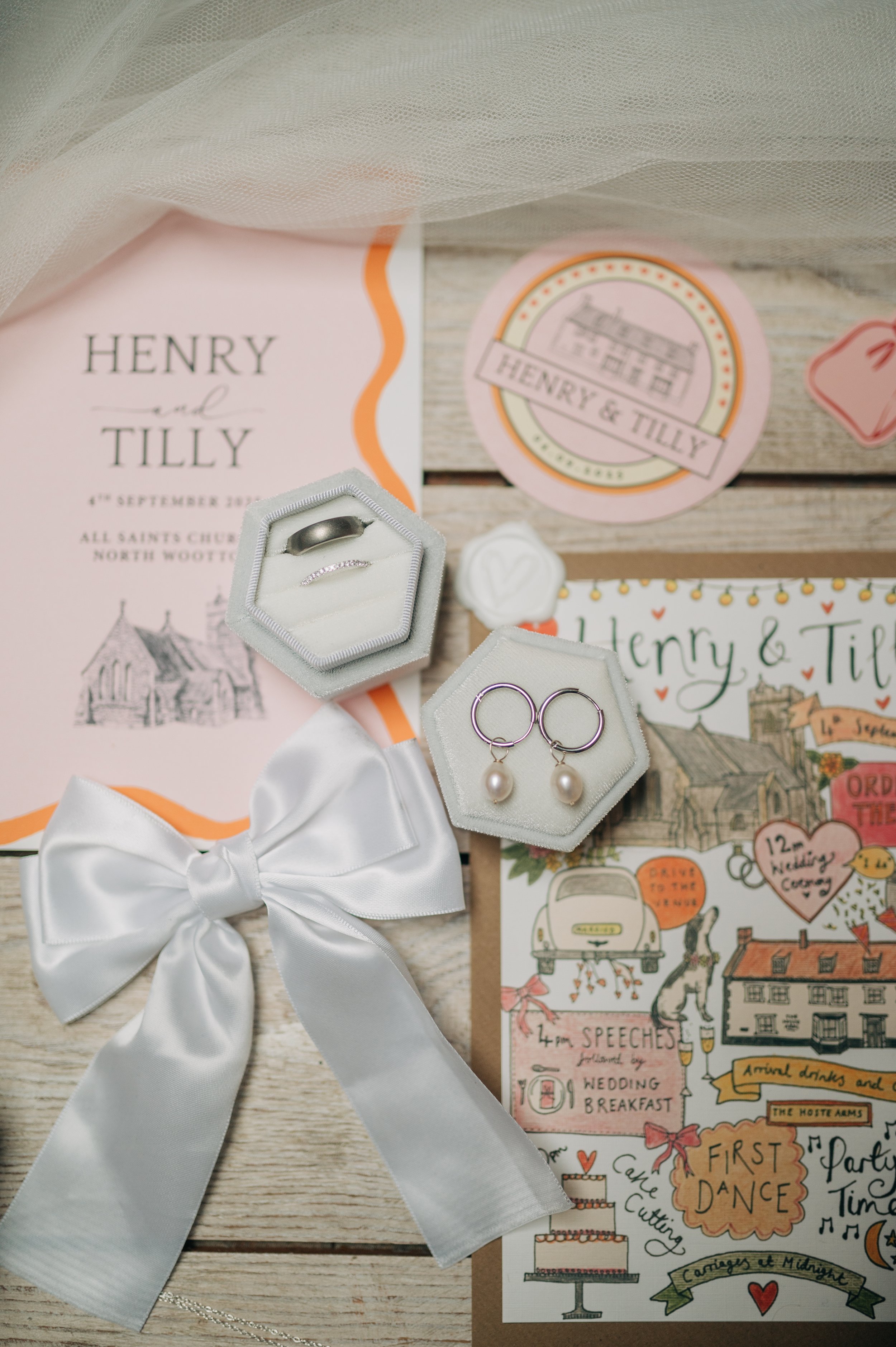 Norfolk Wedding Photographer. Wedding invitation suite with rings in a box, pearl earrings, and a large white satin bow, along with illustrated wedding details and decorations.