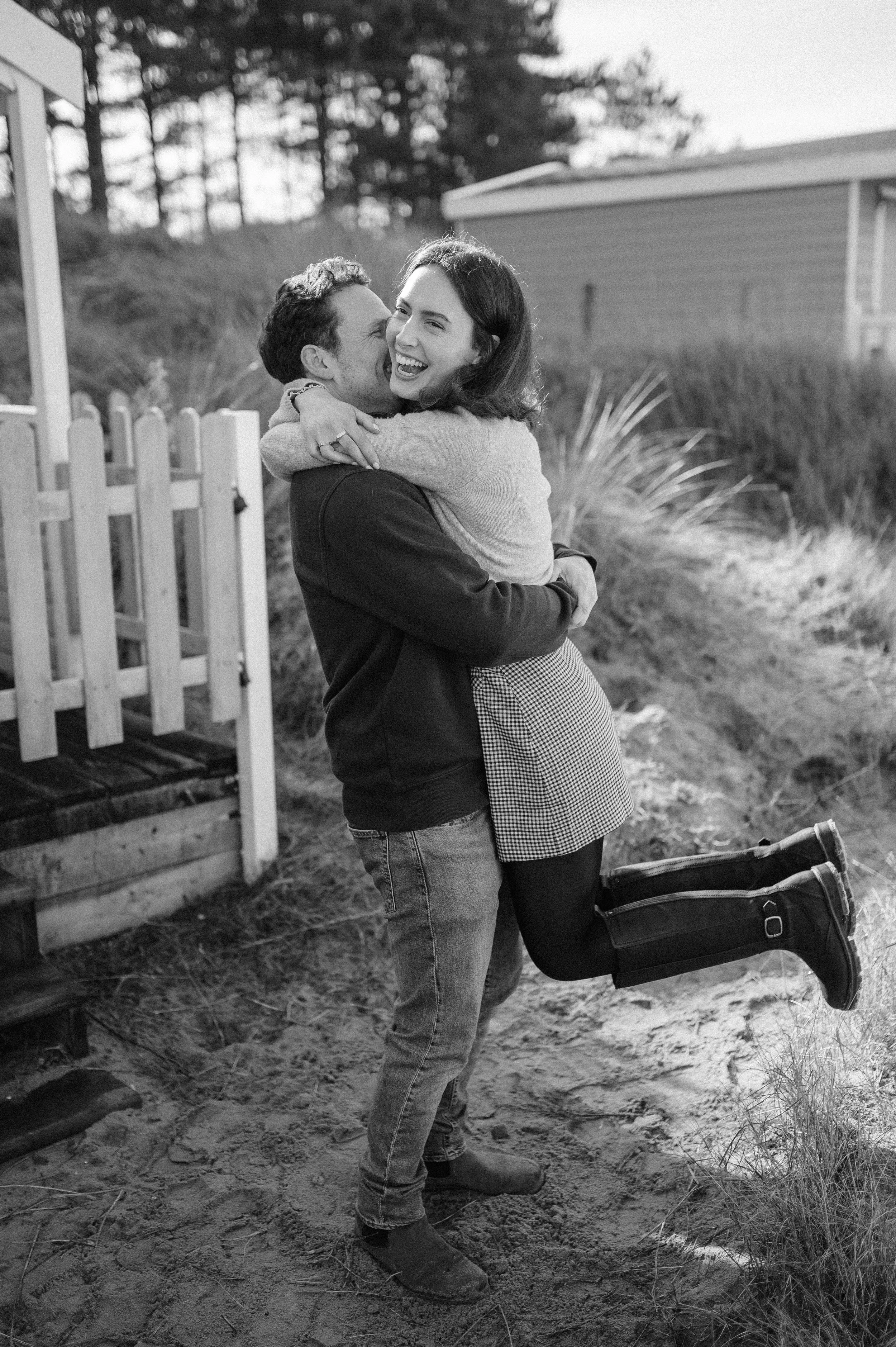 Norfolk Wedding Photographer.  A man and woman sharing an embrace outdoors, woman smiling with one leg raised, trees and a house in the background.