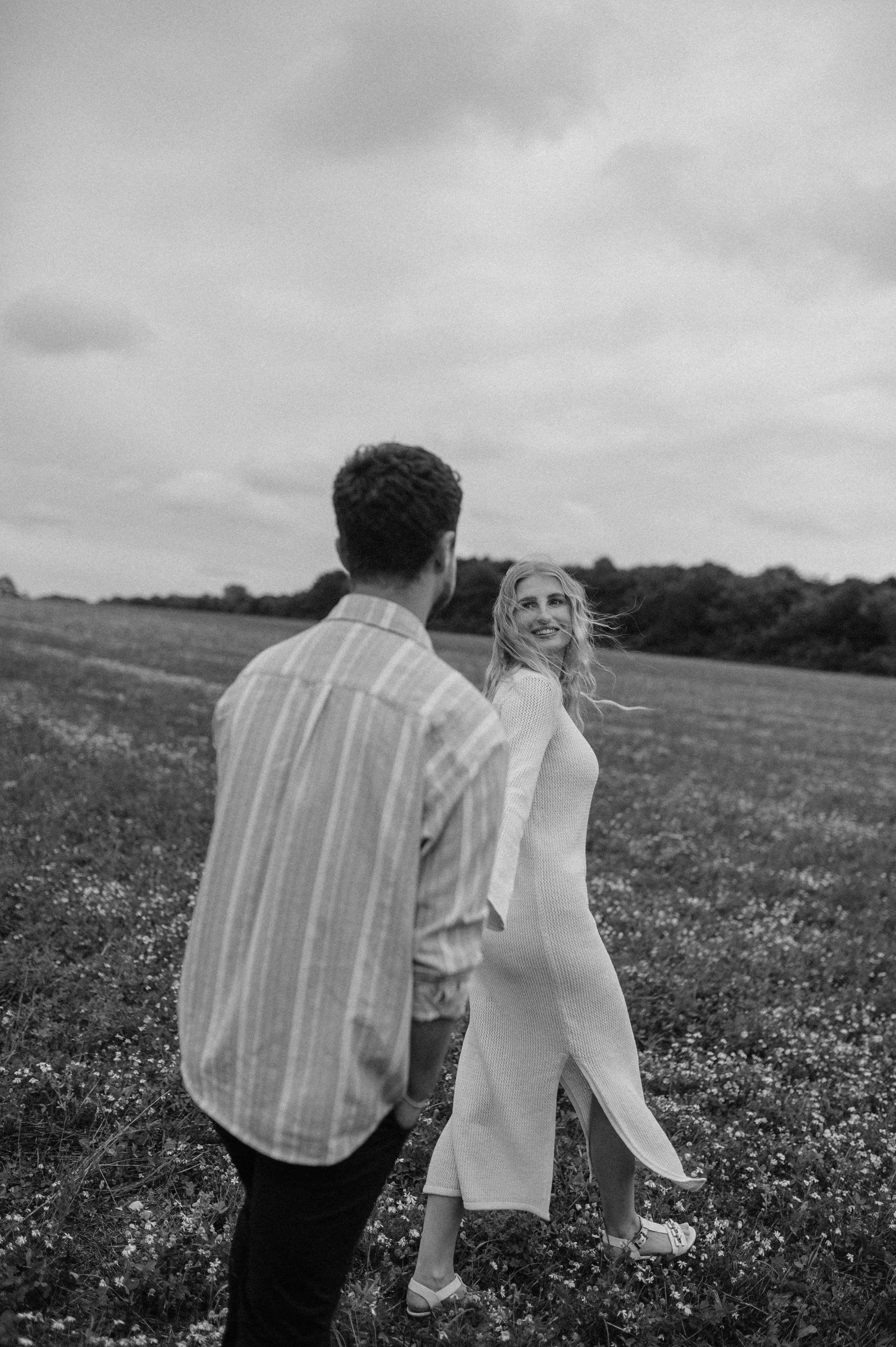 Norfolk Wedding Photographer.  A black-and-white photo of a man and woman walking in a grassy field. The woman is looking back and smiling at the man, who is facing away from the camera. The sky is cloudy.
