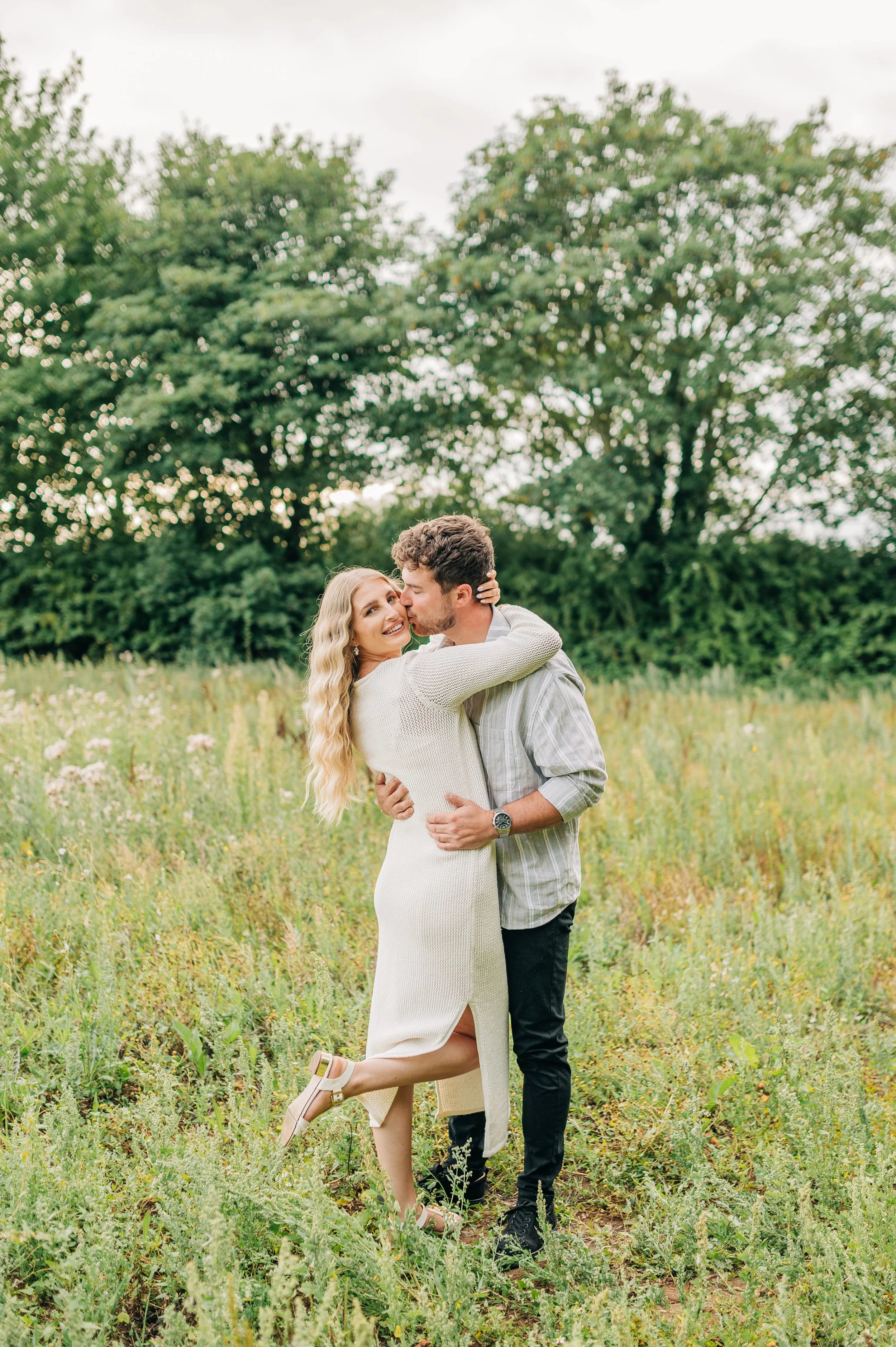 Norfolk Wedding Photographer.  A couple embraces and kisses in a field with green grass and flowers, with trees in the background.