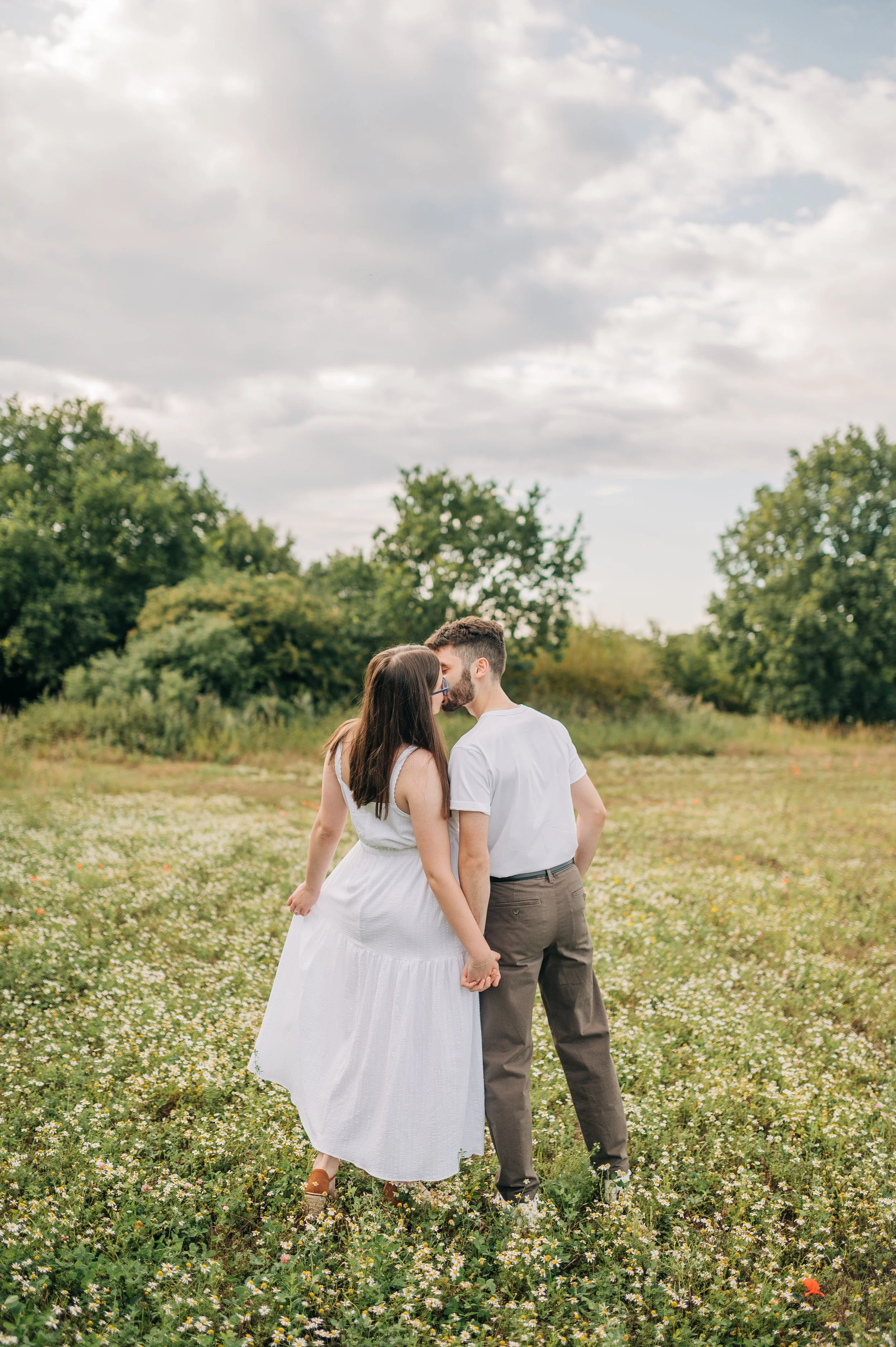 Norfolk Wedding Photographer.  A couple standing in a field holding hands and kissing, with greenery and trees in the background under a cloudy sky.