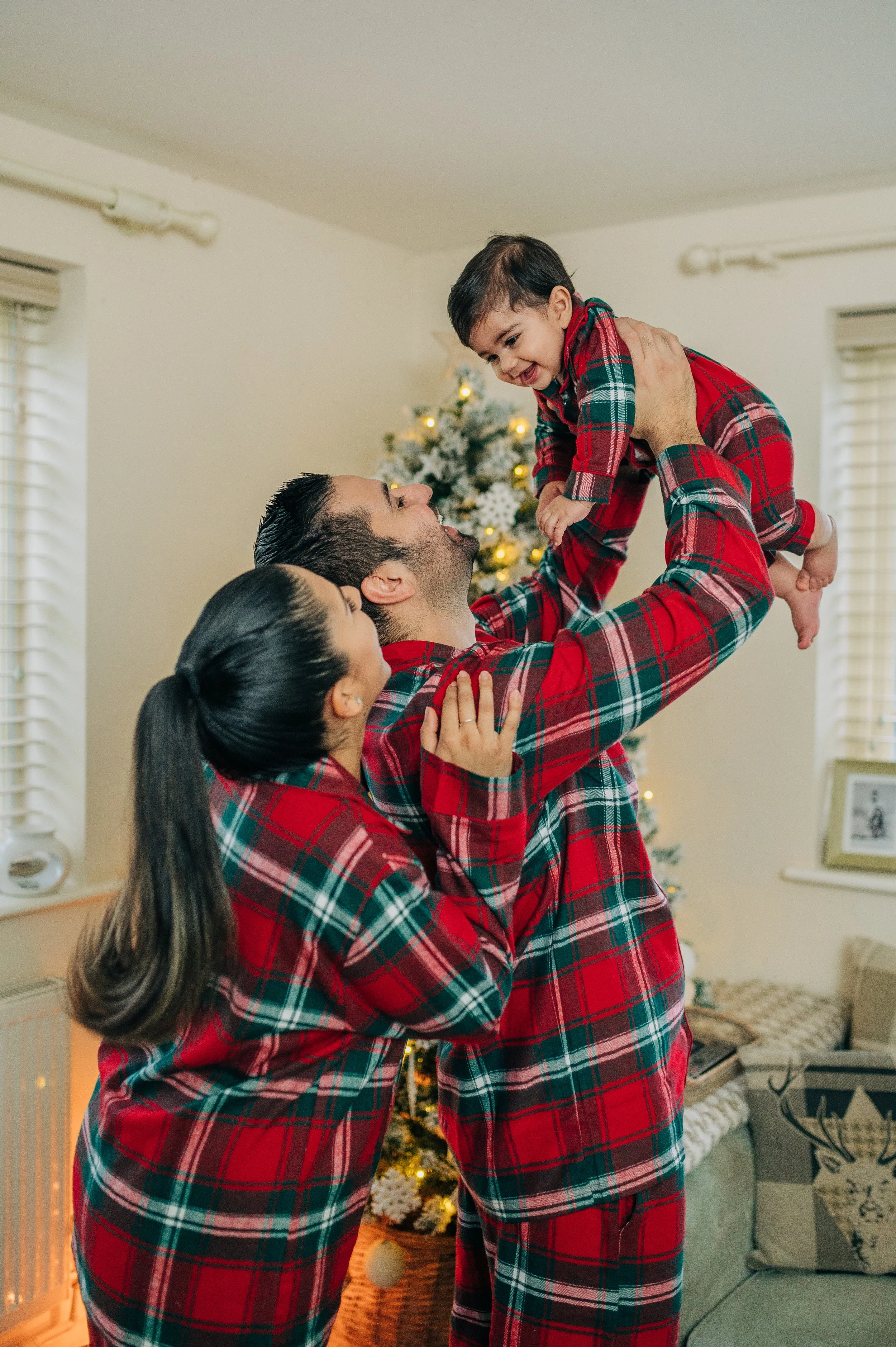 Norfolk Family Photographer.  A family in matching red plaid pajamas celebrating Christmas indoors, with a decorated Christmas tree in the background. The father lifts their smiling young son, while the mother looks on lovingly.