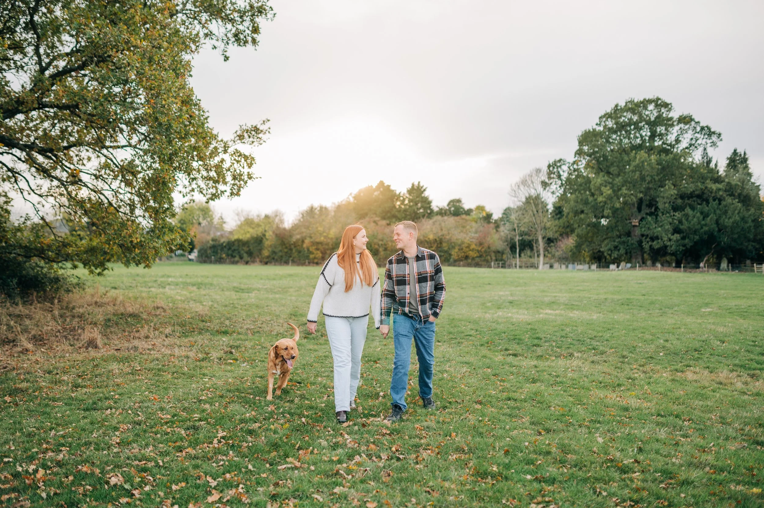 Norfolk Wedding Photographer.  A young couple walking in a grassy park with their dog during fall, with trees and a cloudy sky in the background.