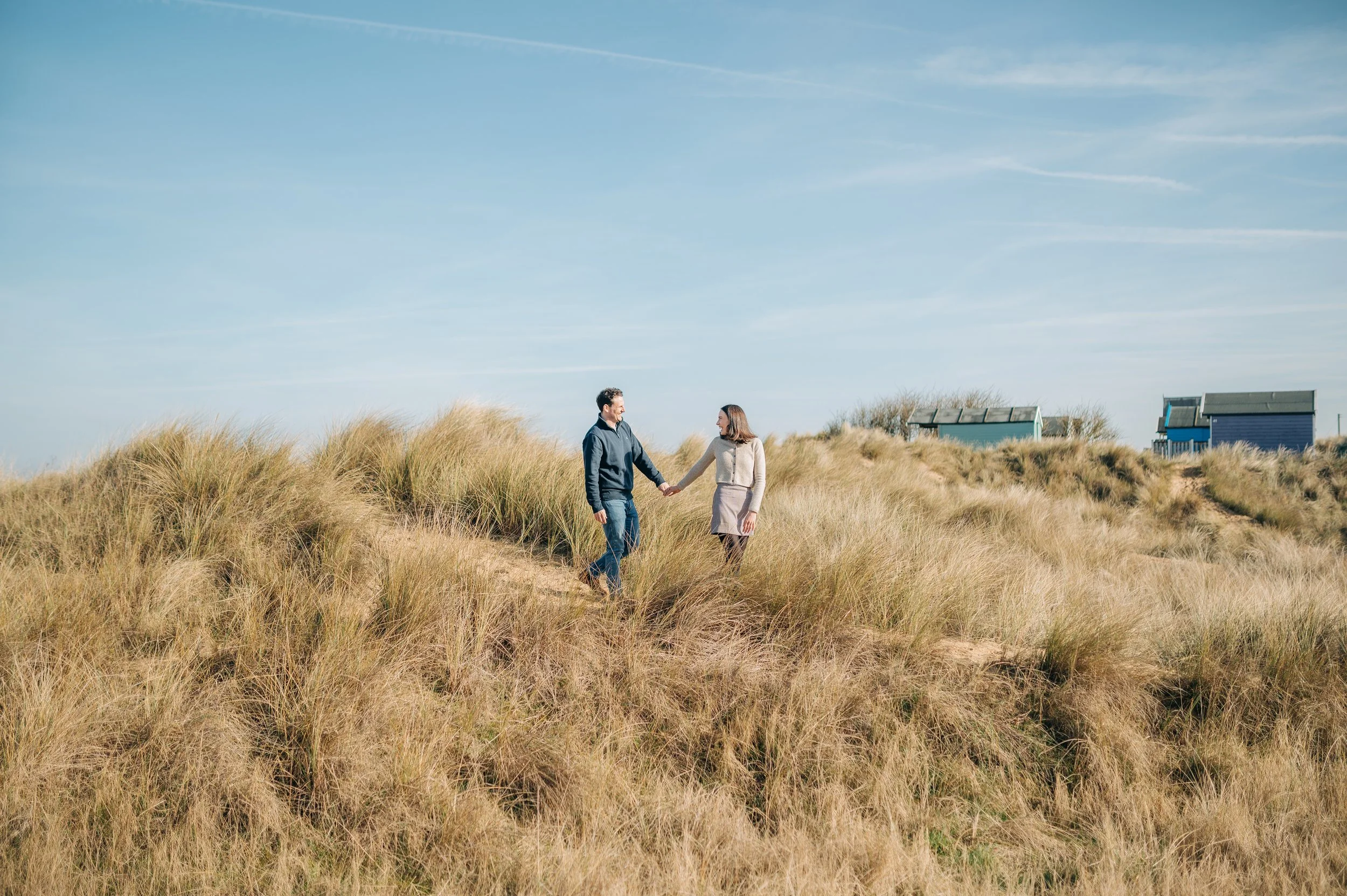 Norfolk Wedding Photographer.  A couple holding hands and walking through a grassy dune area on a clear, sunny day with blue sky and a few clouds, and small beach houses in the background.