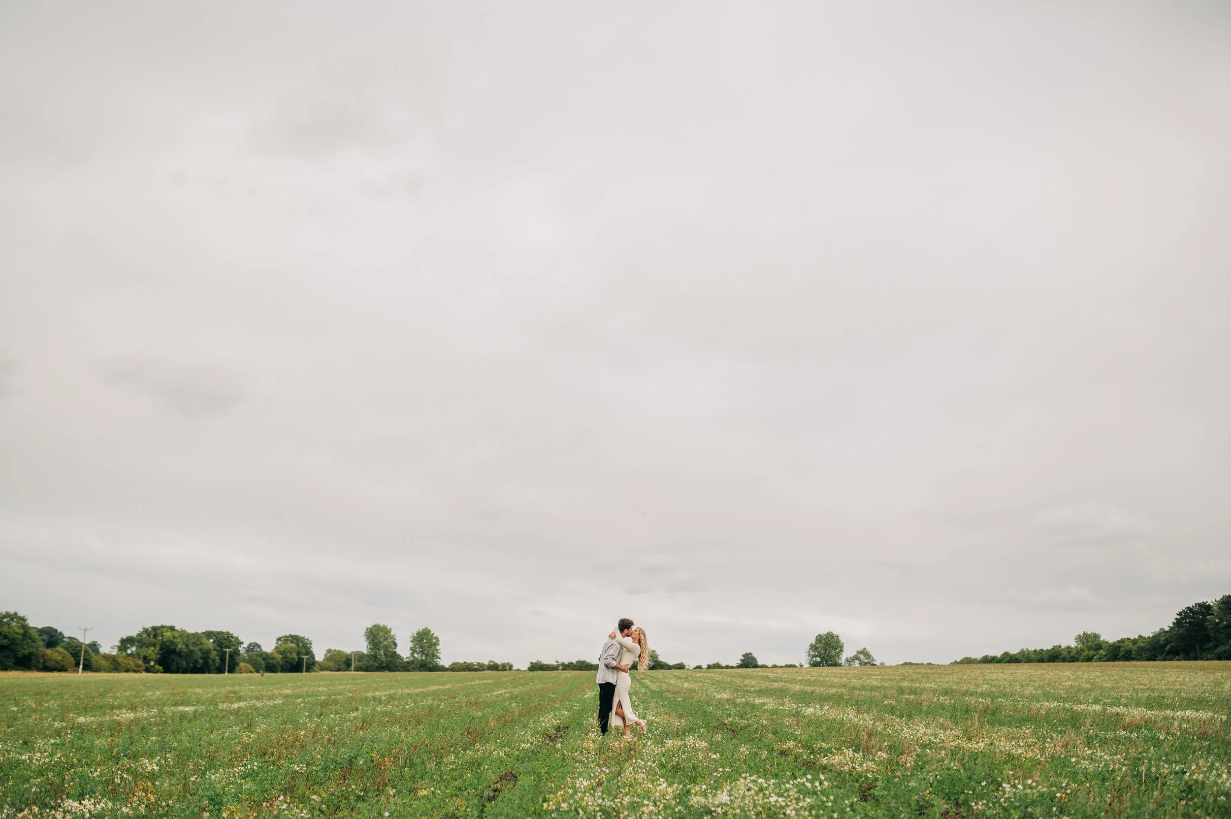 Norfolk Wedding Photographer.  A couple dressed in wedding attire sharing a kiss in the middle of a green field with small white flowers and trees in the background under a cloudy sky.
