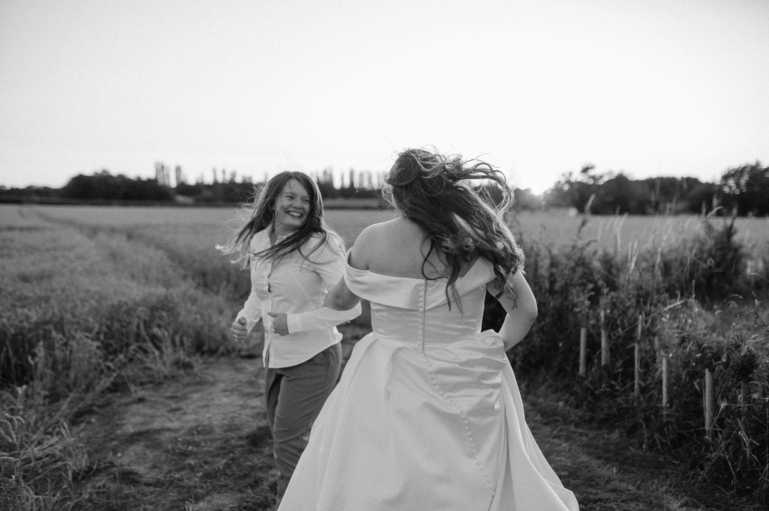 Norfolk Wedding Photographer. Two women are smiling and running in a field, one in a white wedding dress and the other in a white shirt and pants, with wind blowing their hair.