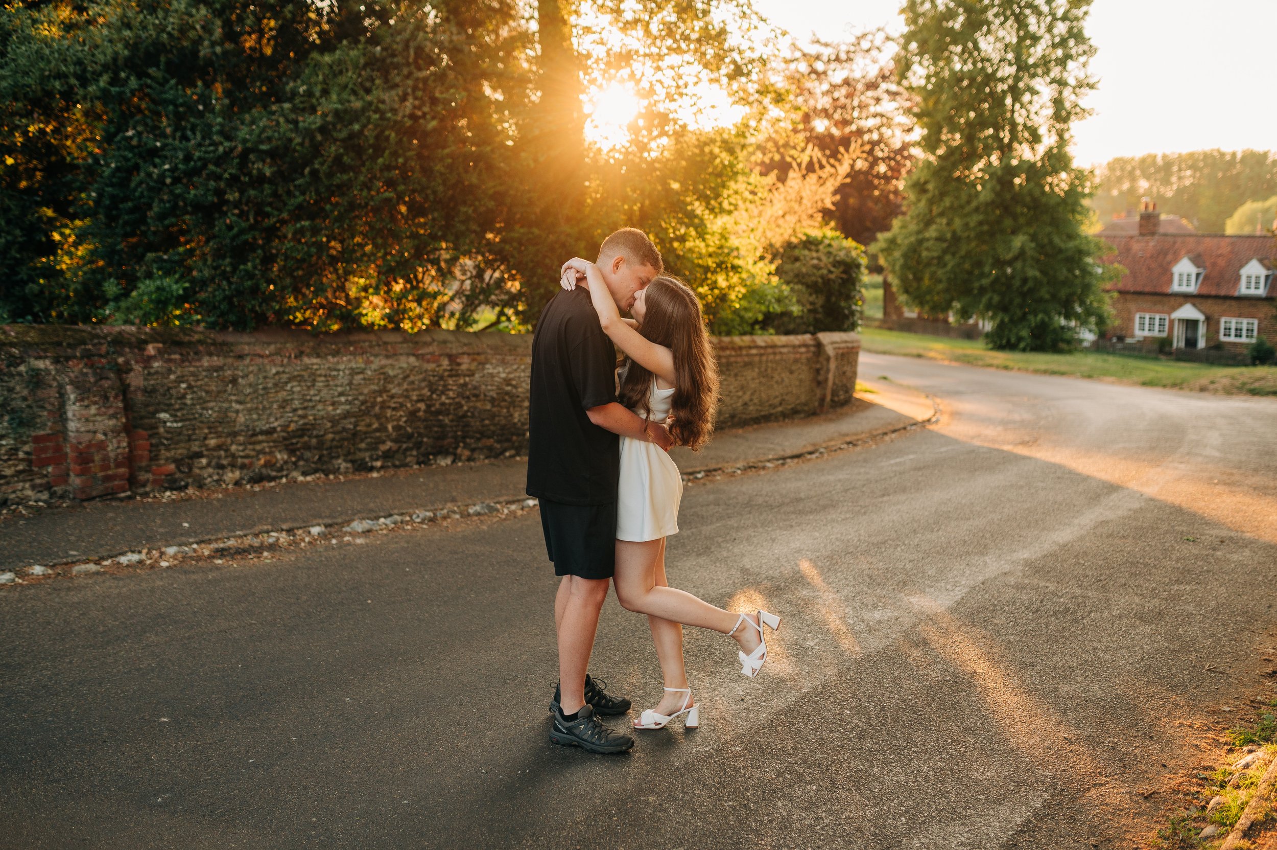 Norfolk Wedding Photographer.  A young man and woman embrace on a street during sunset, with trees and houses in the background.