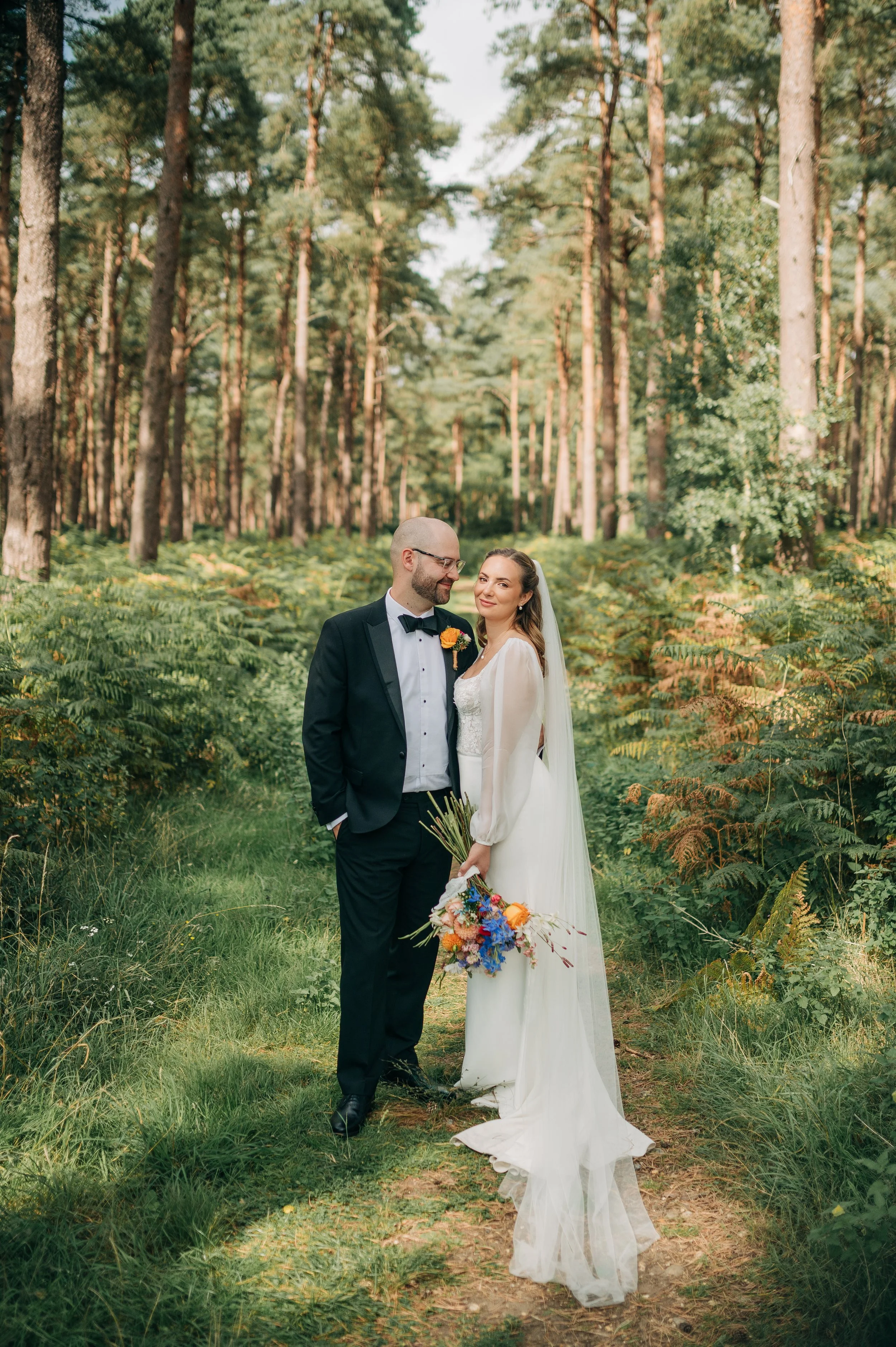 Norfolk Wedding Photographer. A bride and groom standing in a forest, looking at each other, with the bride holding a colorful bouquet.