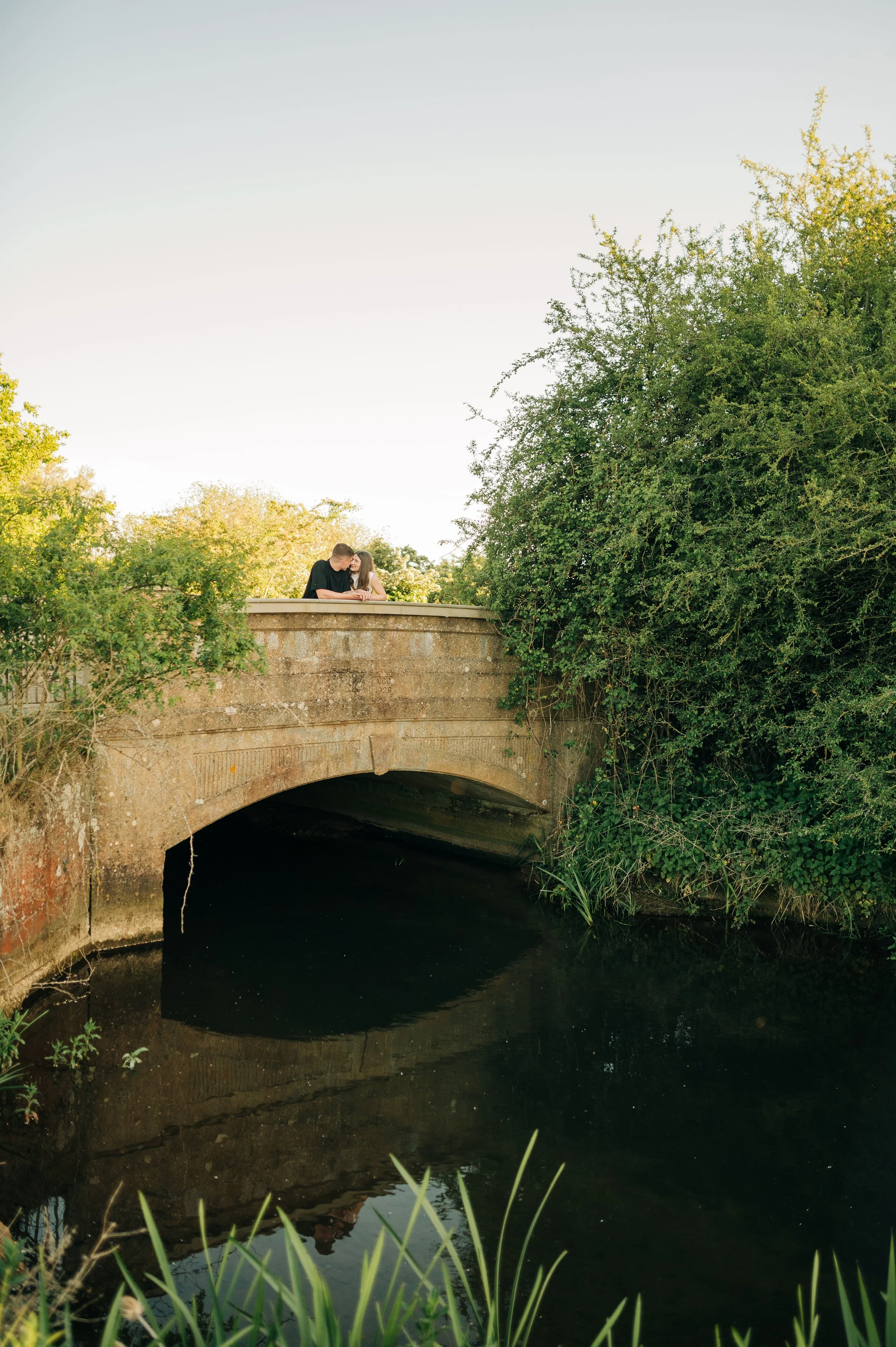 Norfolk Wedding Photographer.  A couple leaning on a stone bridge railing, sharing a kiss, surrounded by greenery under a clear sky.