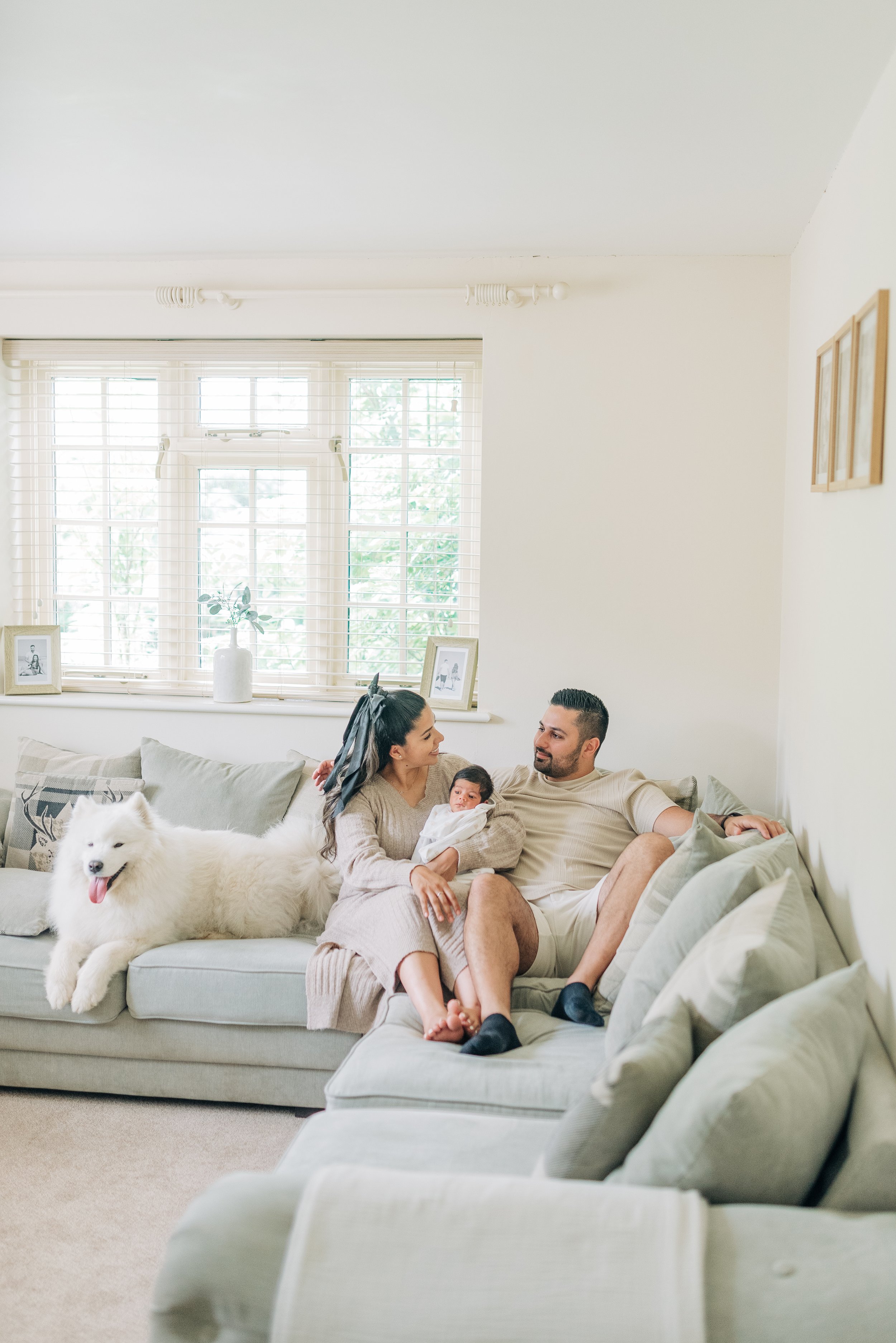 Norfolk Family Photographer.  Family of three with a dog sitting on a beige sofa in a bright living room, holding a newborn baby. The woman and man are smiling at each other, with the dog resting where the woman is sitting.