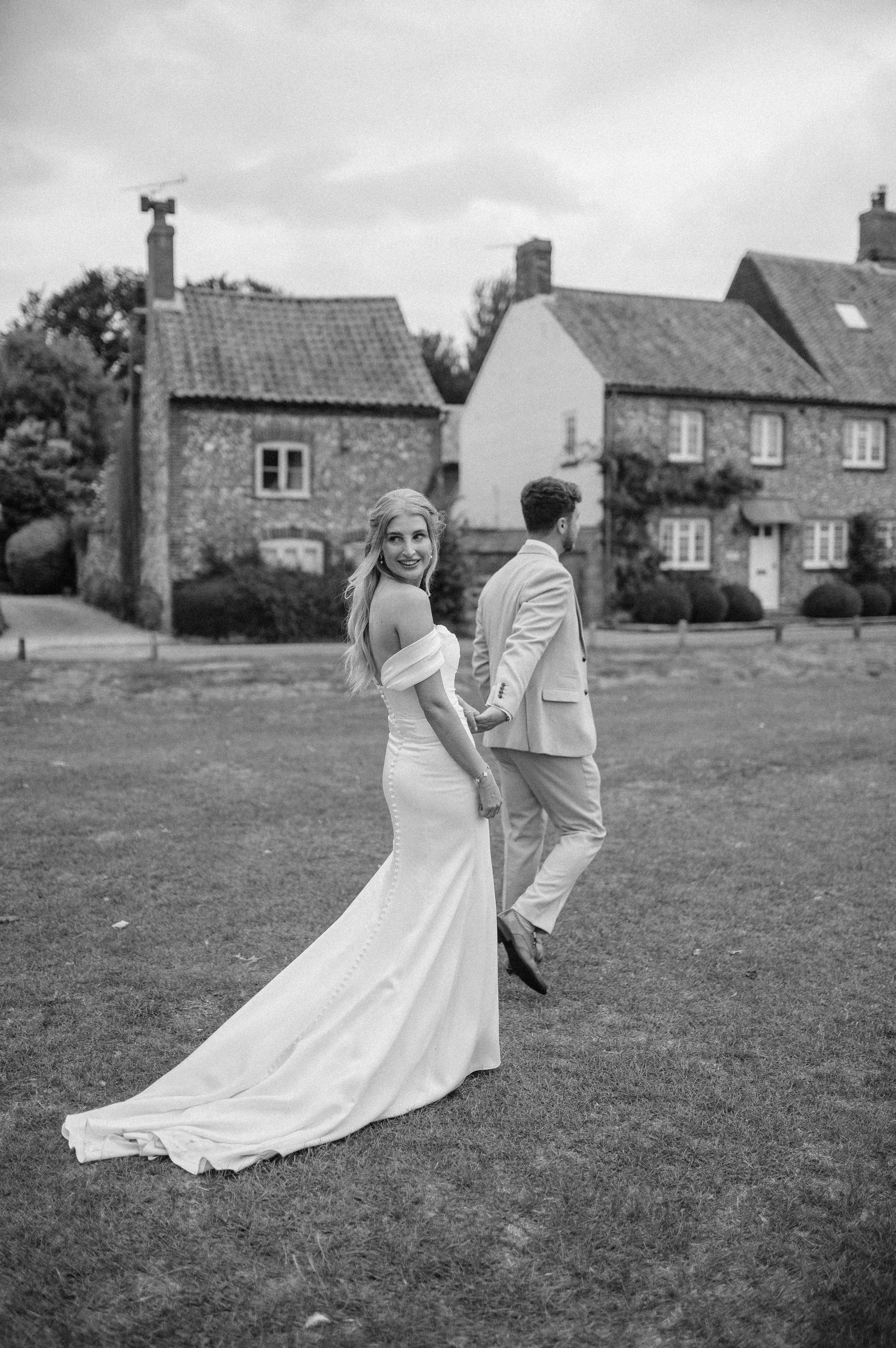 Norfolk Wedding Photographer. Black and white photo of a bride in a long wedding gown with off-shoulder sleeves and a groom in a suit holding hands and walking on a grassy field in front of a row of houses.