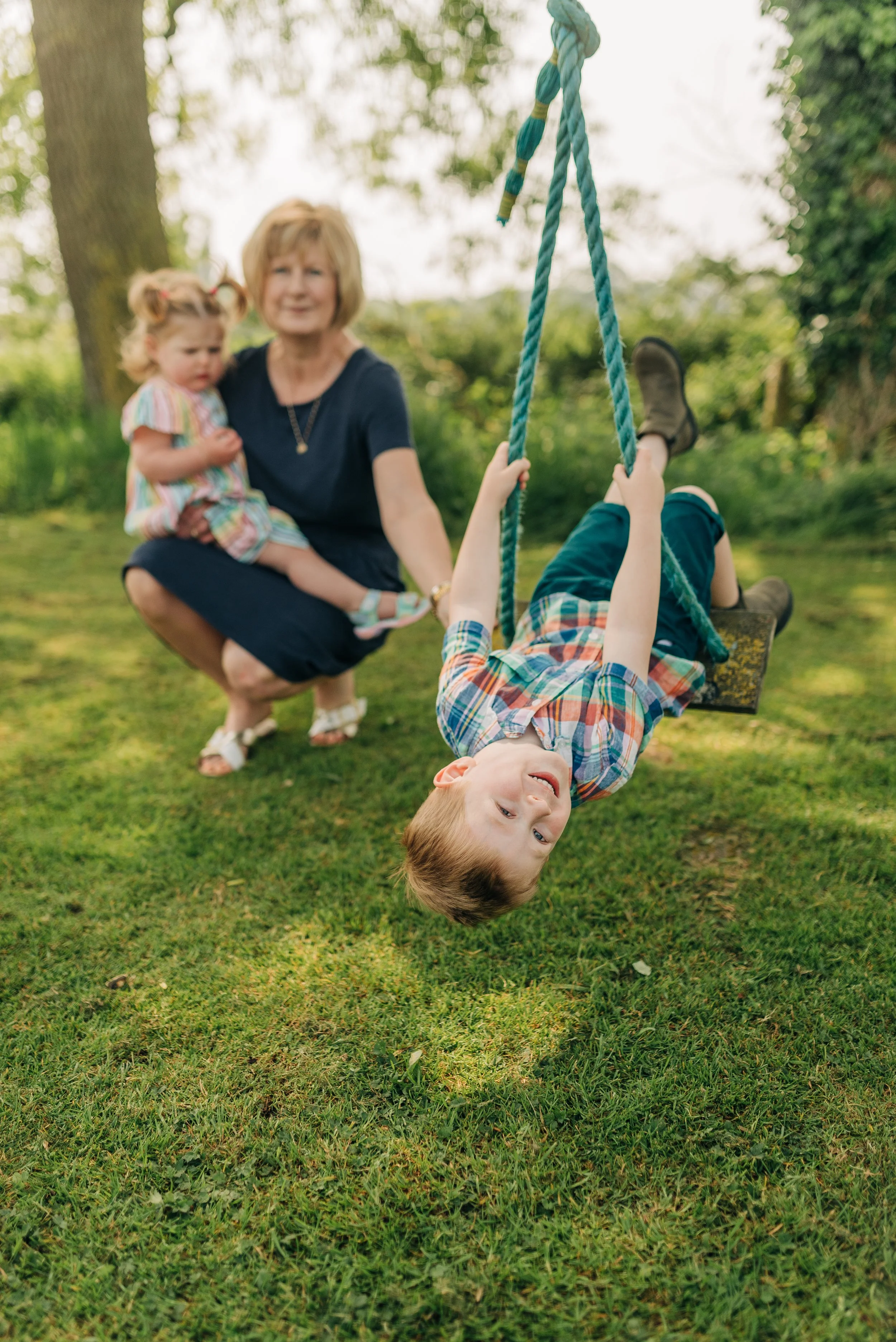 Norfolk Family Photographer.  A young boy is swinging upside down on a rope swing in a grassy park, while a woman and a young girl watch in the background.