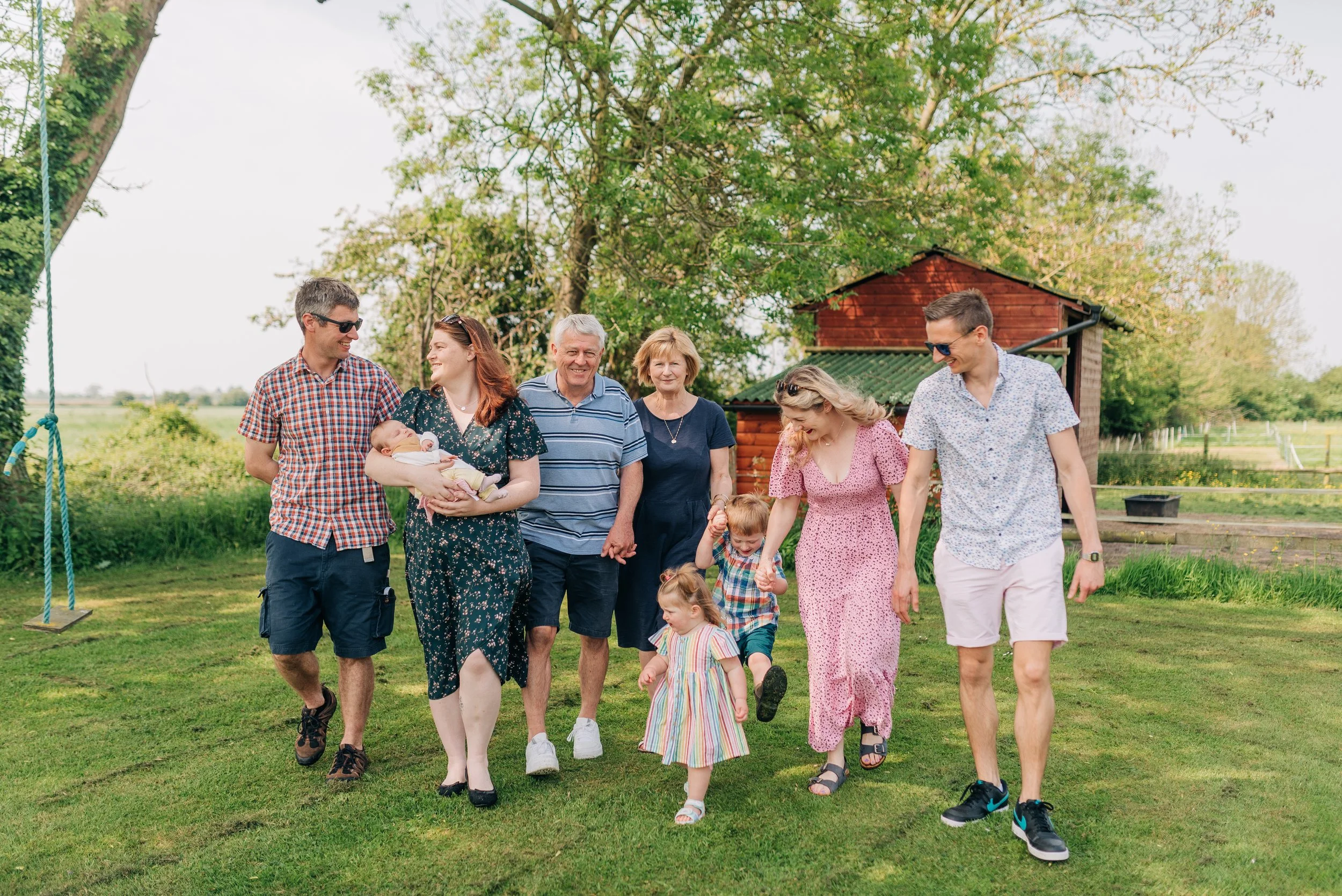 Norfolk Family Photographer.  A multi-generational family walking together outdoors in a green yard with trees and a red wooden shed, celebrating.