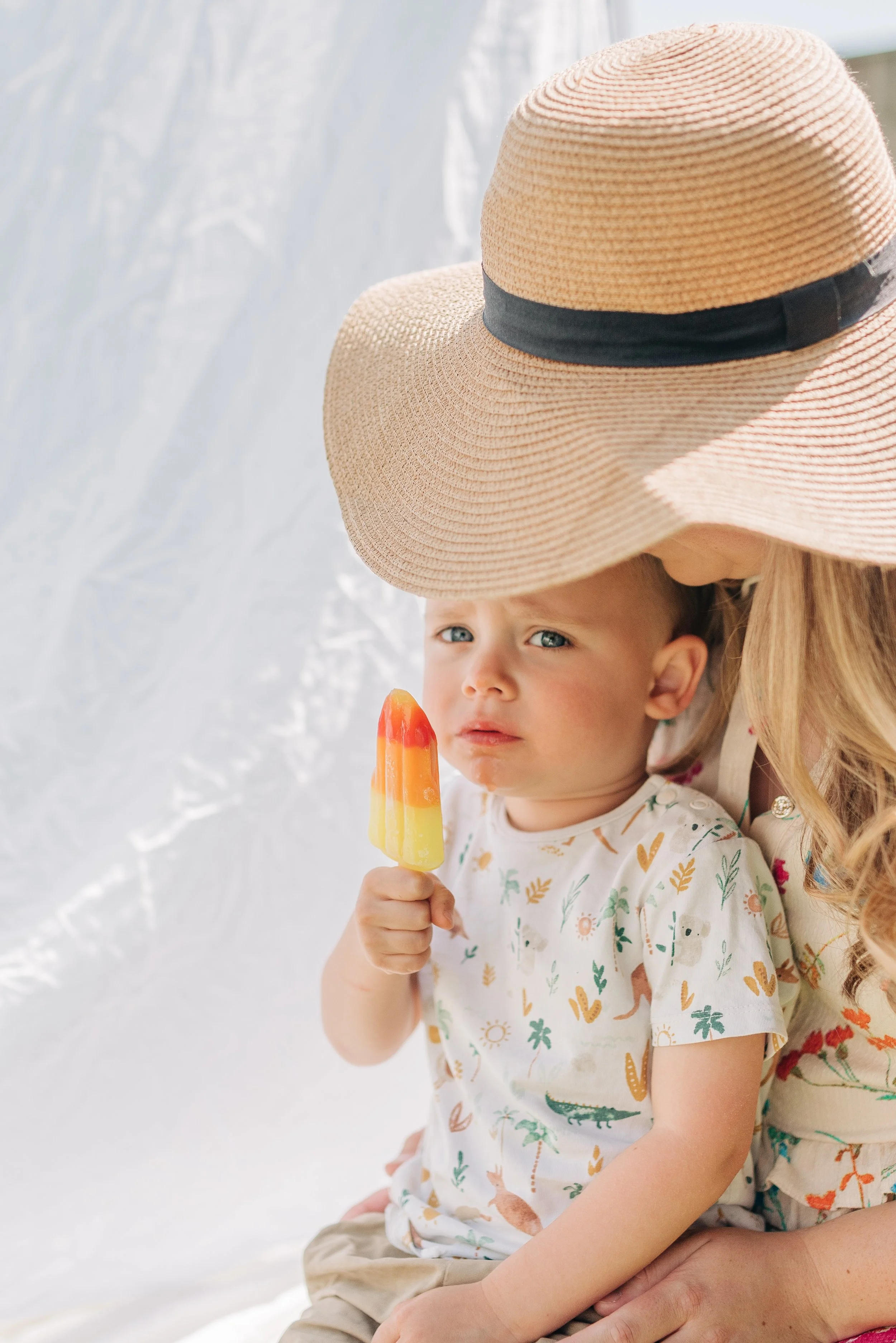 Norfolk Family Photographer.  A woman wearing a large sunhat sitting with a young boy holding a multicolored popsicle, both on a sunny day near a white background.