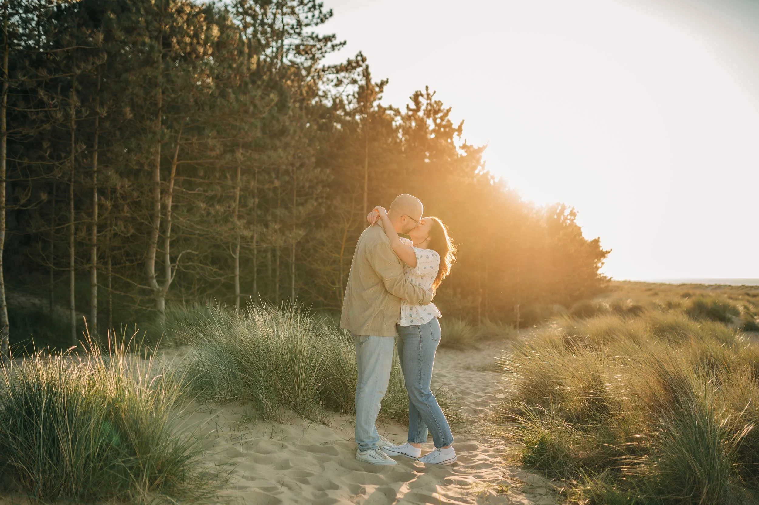 Norfolk Wedding Photographer.  A couple kissing on a sandy path surrounded by tall grass with trees in the background at sunset.
