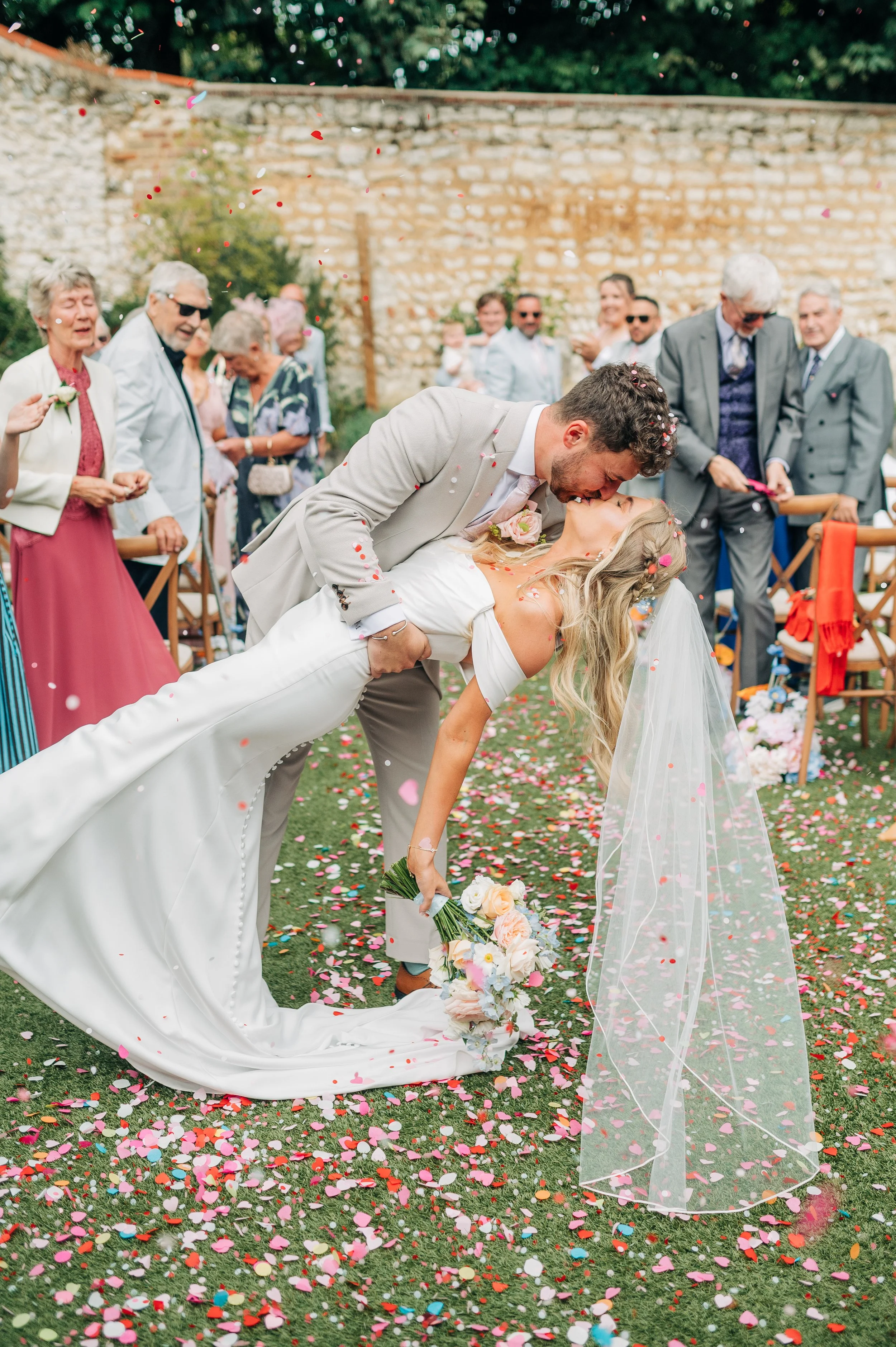 Norfolk Wedding Photographer. A newlywed couple sharing a kiss during their outdoor wedding celebration, with guests watching and colorful confetti on the ground.