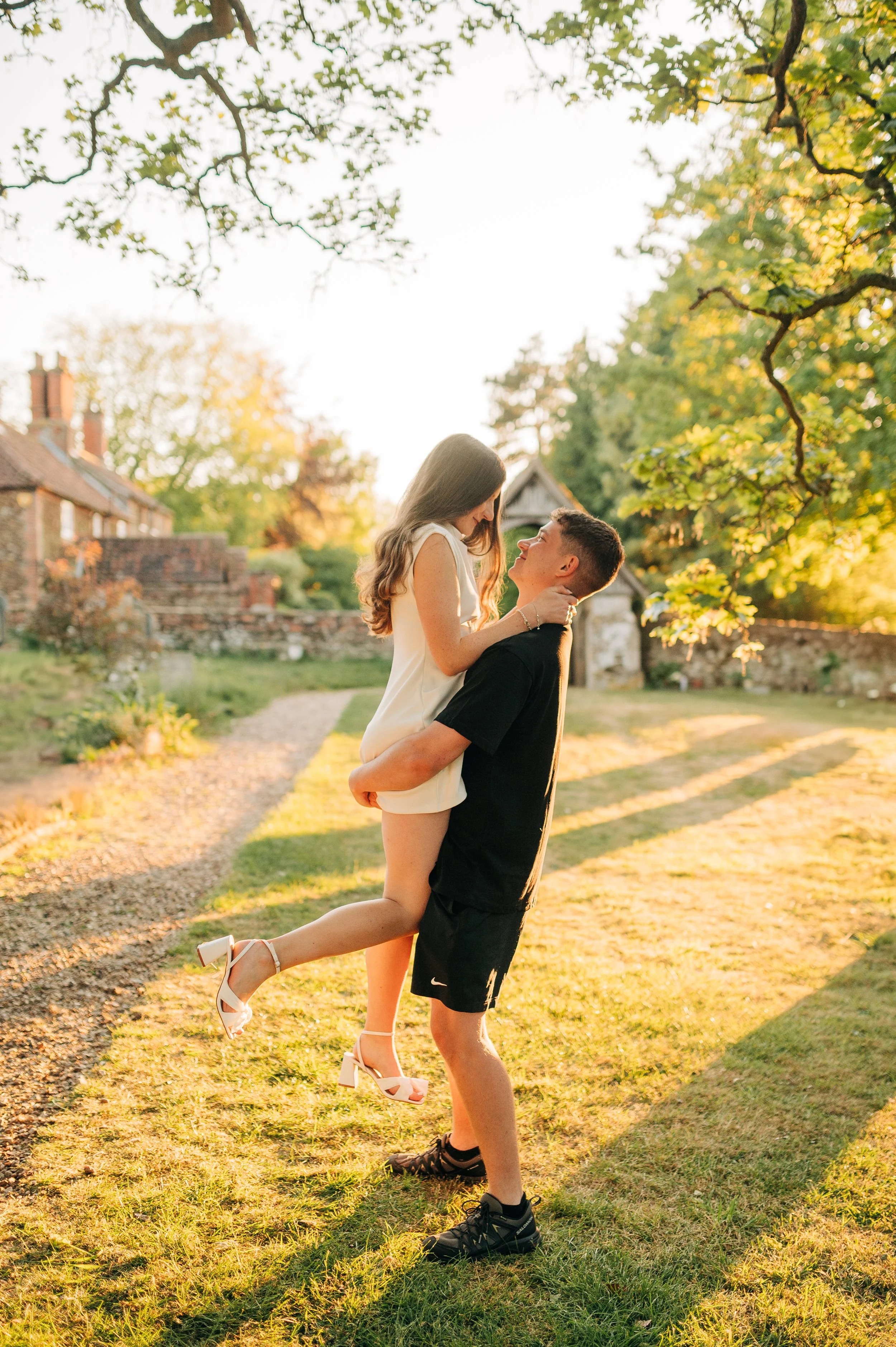 Norfolk Wedding Photographer.  A young couple outdoors during sunset, with the man lifting the woman in his arms, both smiling at each other. The woman is wearing a white dress and heels, and the man is dressed in black casual clothes.